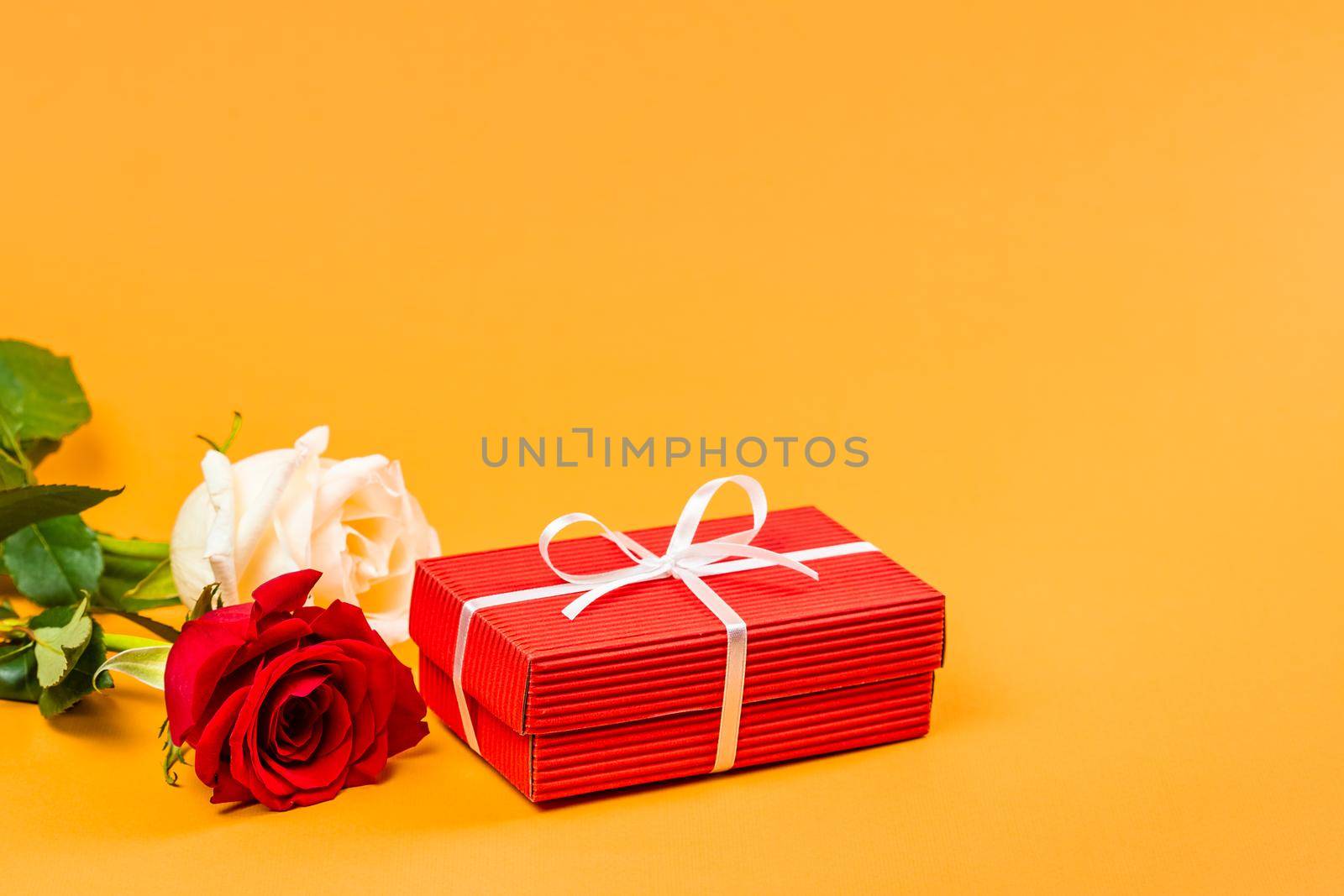 Red gift box with a white ribbon and two red and white roses over vibrant orange background. Valentines Day or Birthday gift concept 