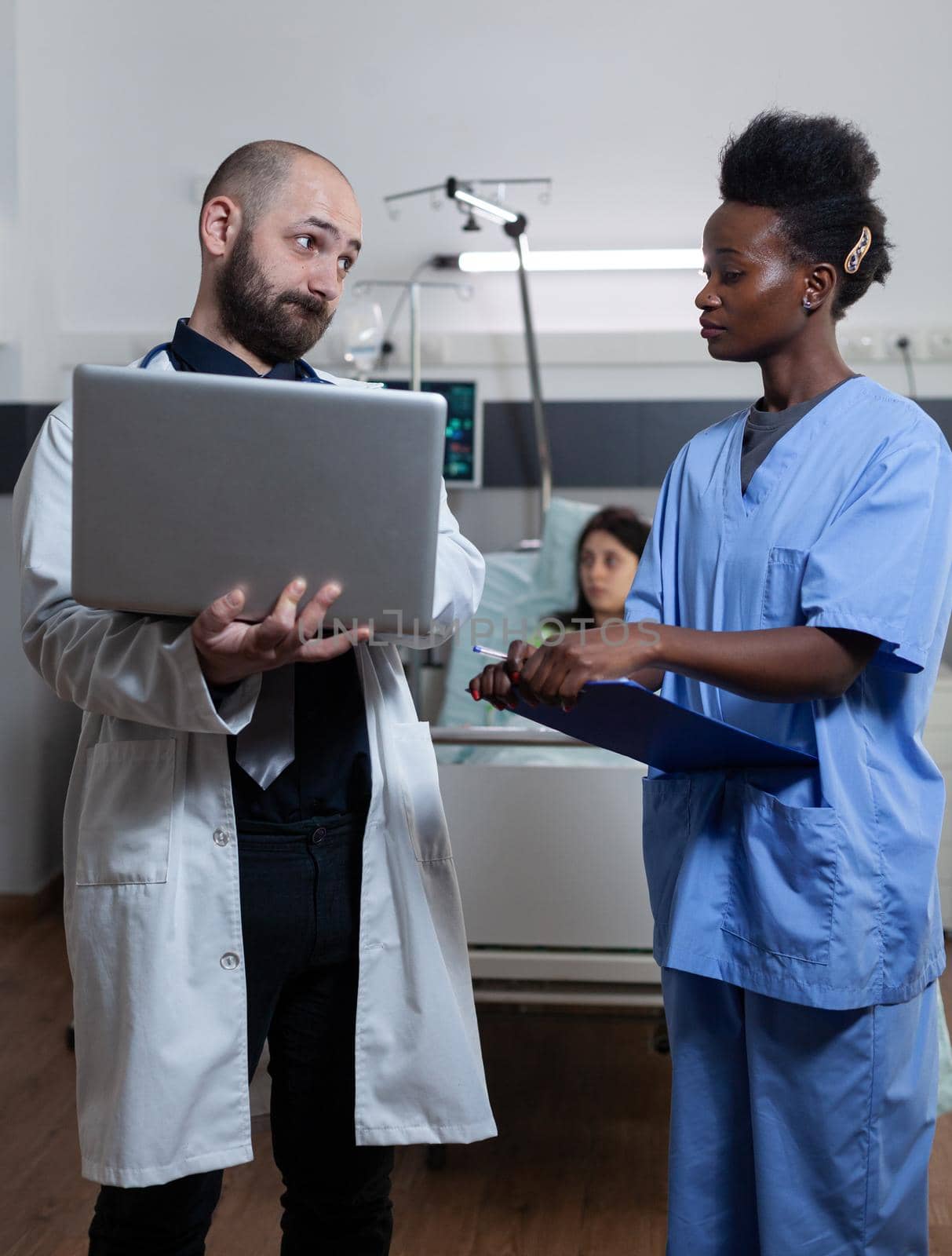 Doctor holding laptop unhappy with patient lab results looking with disapproval at nurse holding clipboard in hospital ward. Physician discussing medical treatment with health care worker.