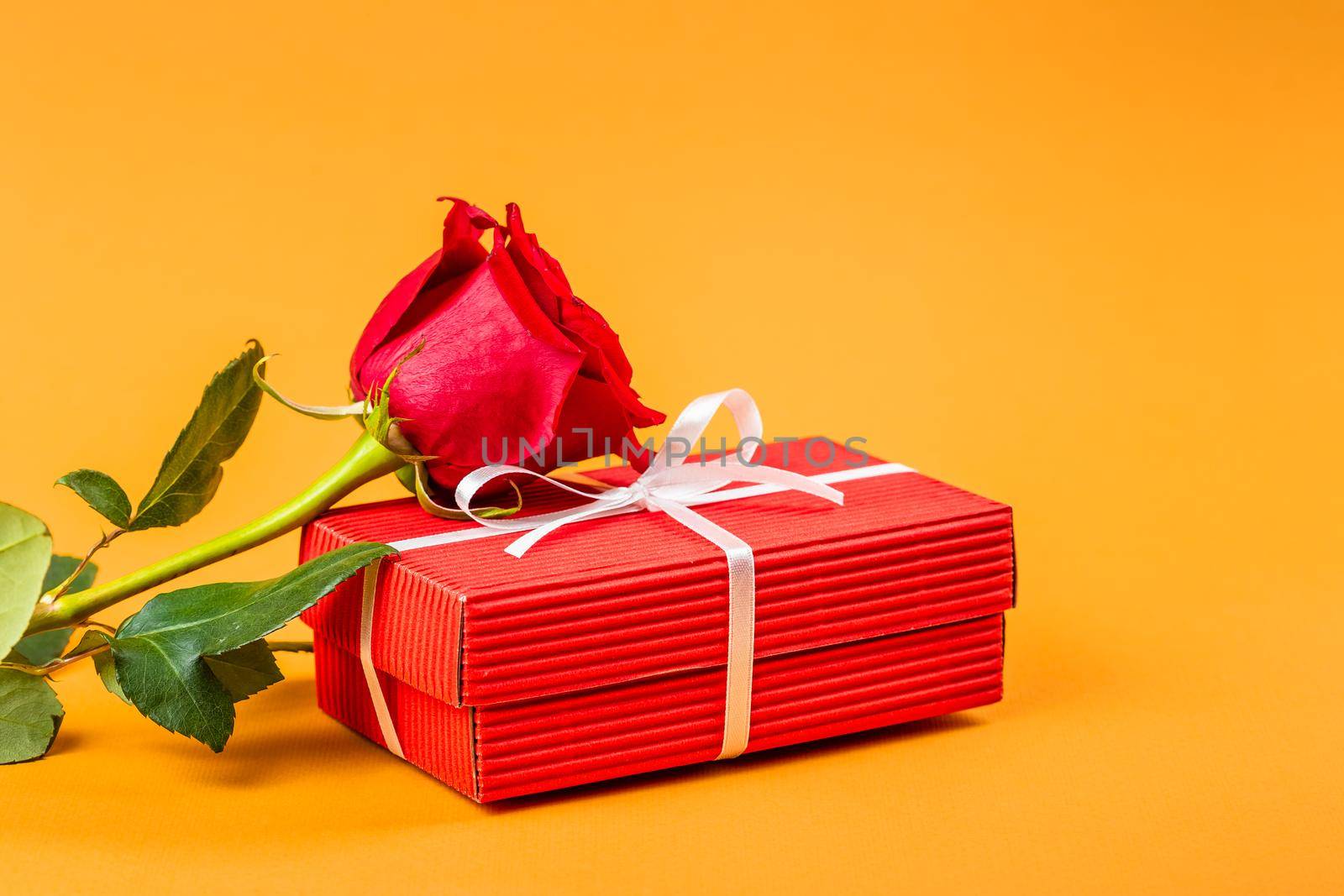 Red gift box with a white ribbon and white rose flower over vibrant orange background. Valentines Day or Birthday gift concept 