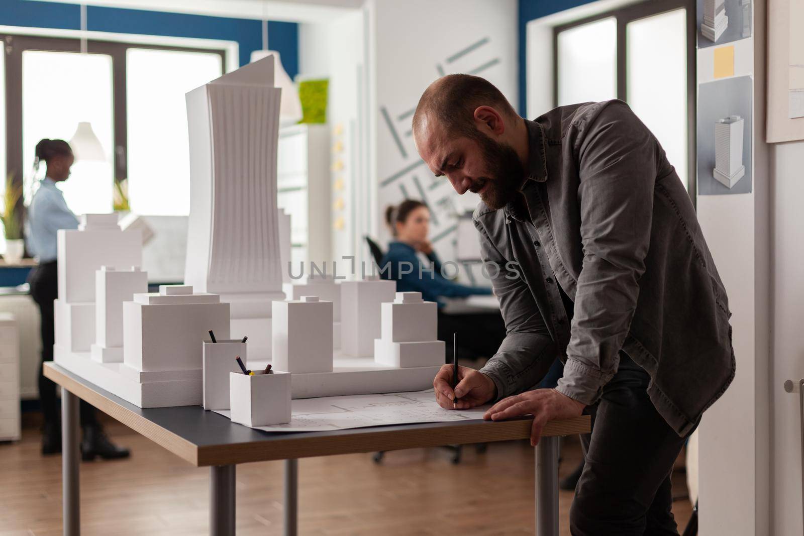 Architect drawing blueprints at desk in modern architectural office next to white foam scale model of skyscraper. Engineer making notes on construction plan leaning over desk.