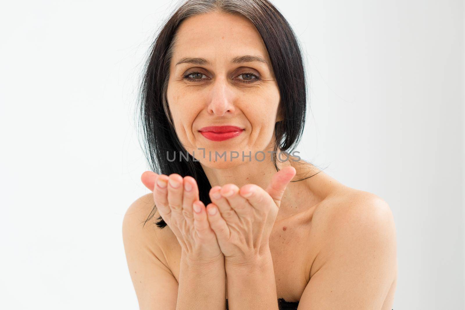 Middle aged woman starting getting grey-haired is sending air kiss in studio with naked shoulders on white background, face skin care beauty, middle age skincare cosmetics, cosmetology concept.