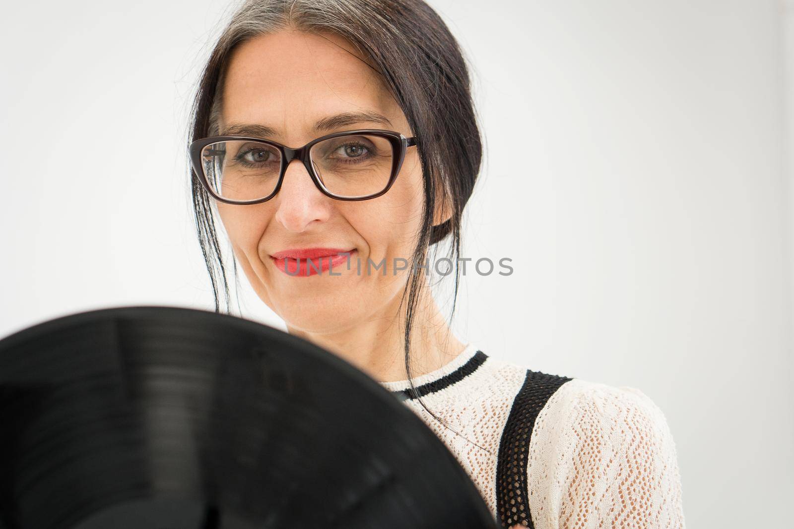 Studio photo of middle aged woman starting getting grey-haired wearing black and white clothes with vinyl record in hands on white background, middle age sexy lady, happy life concept by balinska_lv