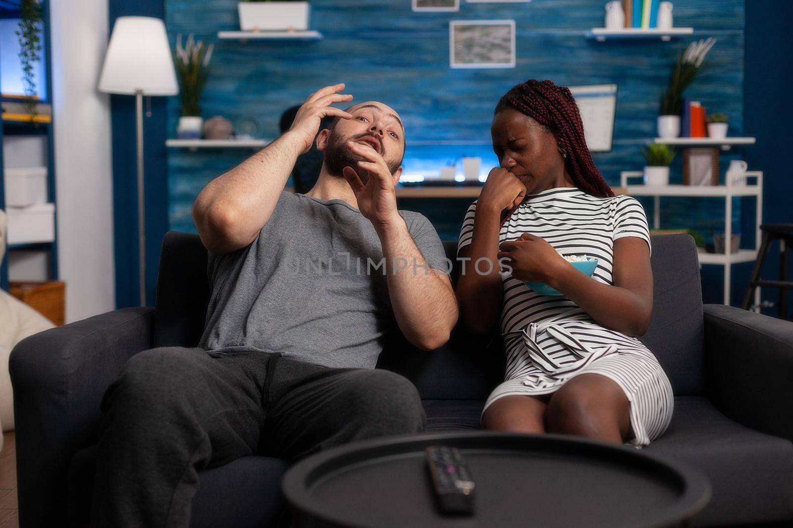 Scared man and woman watching horror film on television. Terrified interracial couple sitting on the sofa while enjoying scary movies. Multiethnic friends watching spooky show and eating popcorn at home