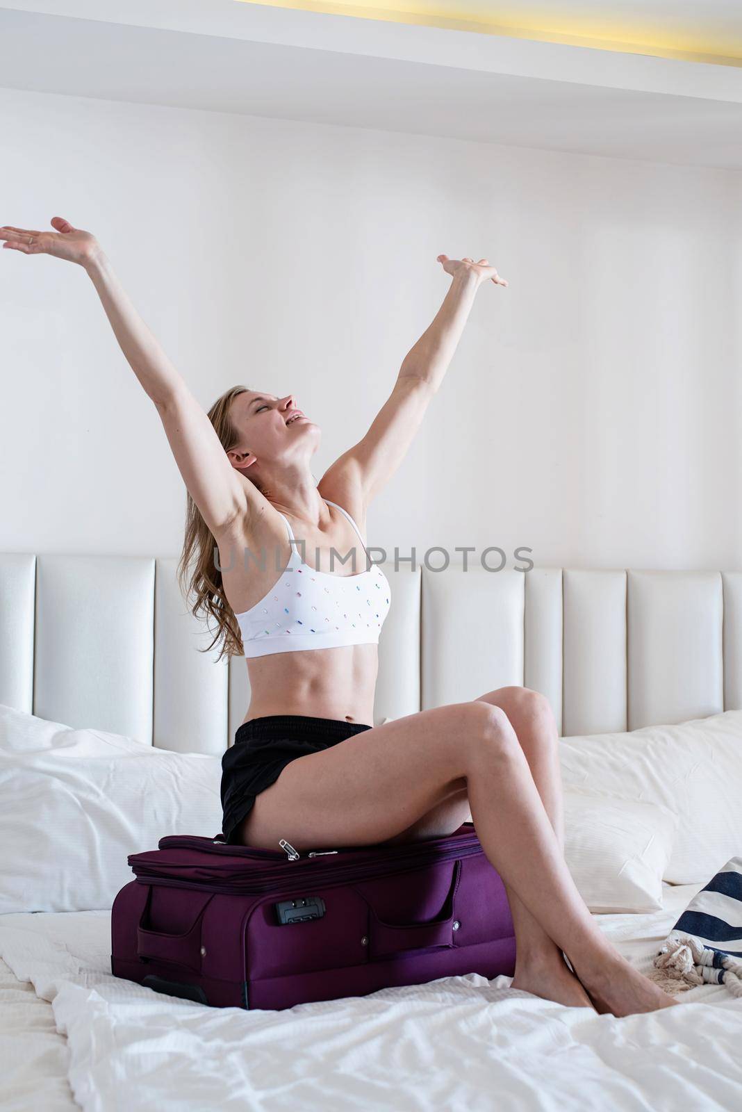 Excited young caucasian woman in swimsuit packing bags for holidays, pretty woman preparing suitcase for vacations getting ready for travel, sitting on bags