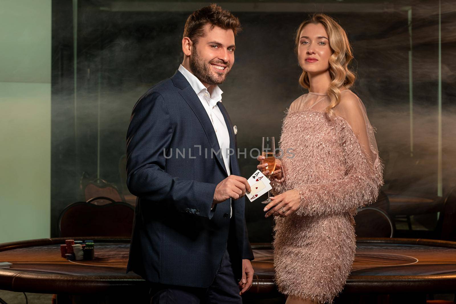 Couple of successful poker players standing near gaming table with stack of betting chips on background of smoky interior of empty casino hall. Confident woman champagne, smiling man showing two aces