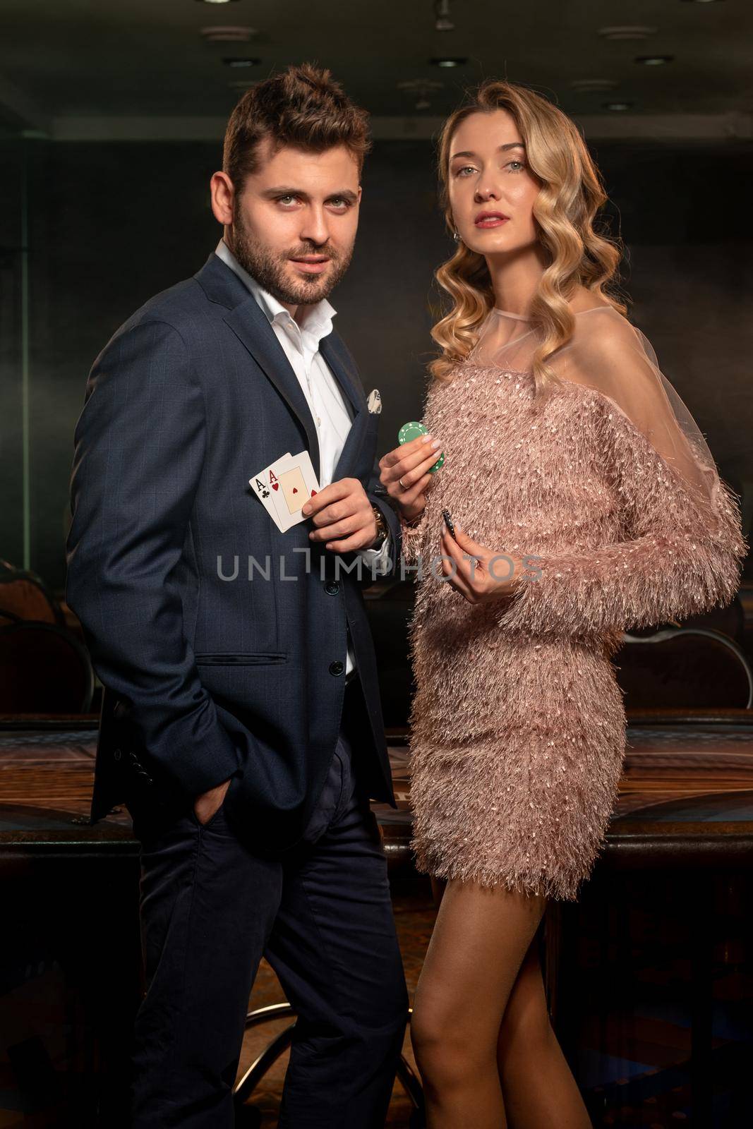 Portrait of successful poker players. Proud young bearded man and attractive blonde girl winning in casino, standing in front of gaming table with cards and betting chips in hands. Gambling concept