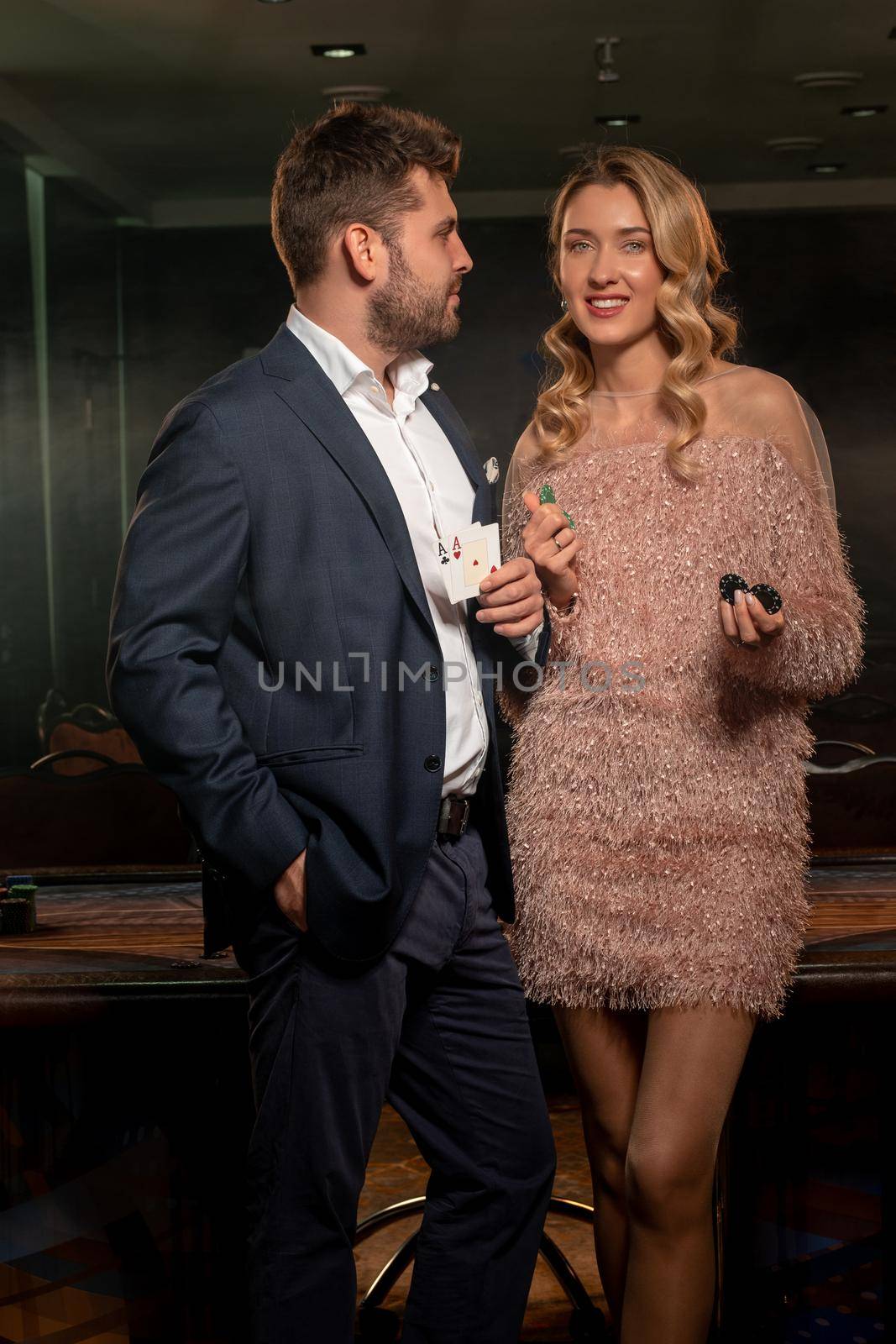 Lucky young blonde woman and bearded guy winning at poker, standing in front of gaming table in casino, holding cards and betting chips. Successful gambling concept