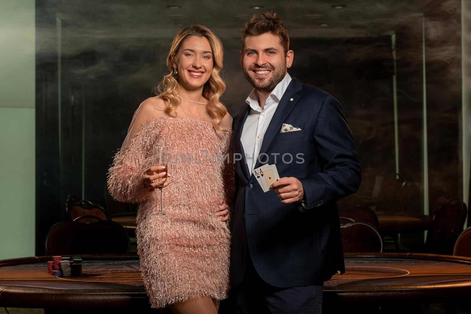 Happy young elegant couple posing in casino near poker table. Smiling woman holding glass of champagne, cheerful man showing winning card set of pair of aces. Gambling concept