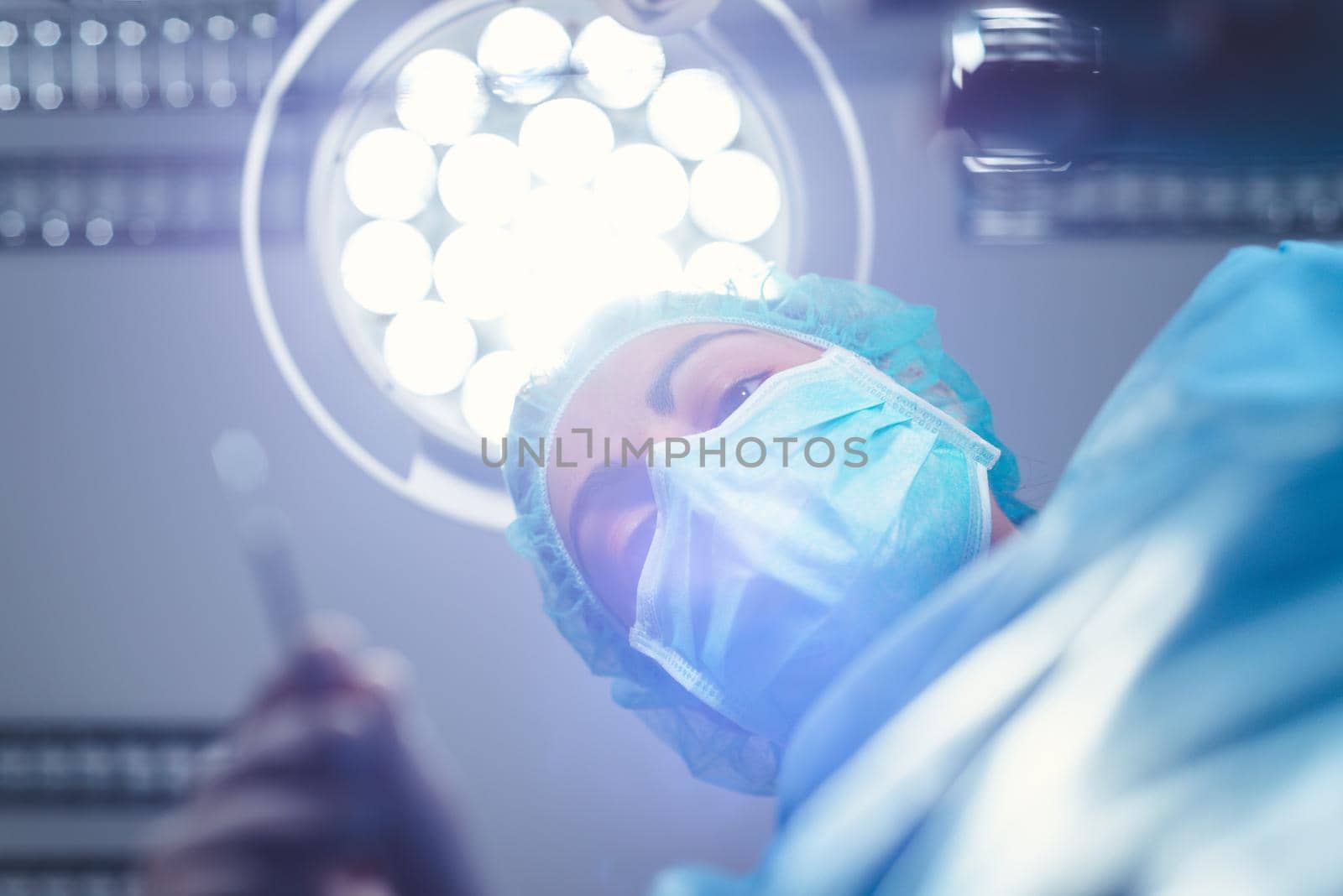 From below woman surgeon in medical uniform using professional tools while standing under bright light in operating theater by HERRAEZ
