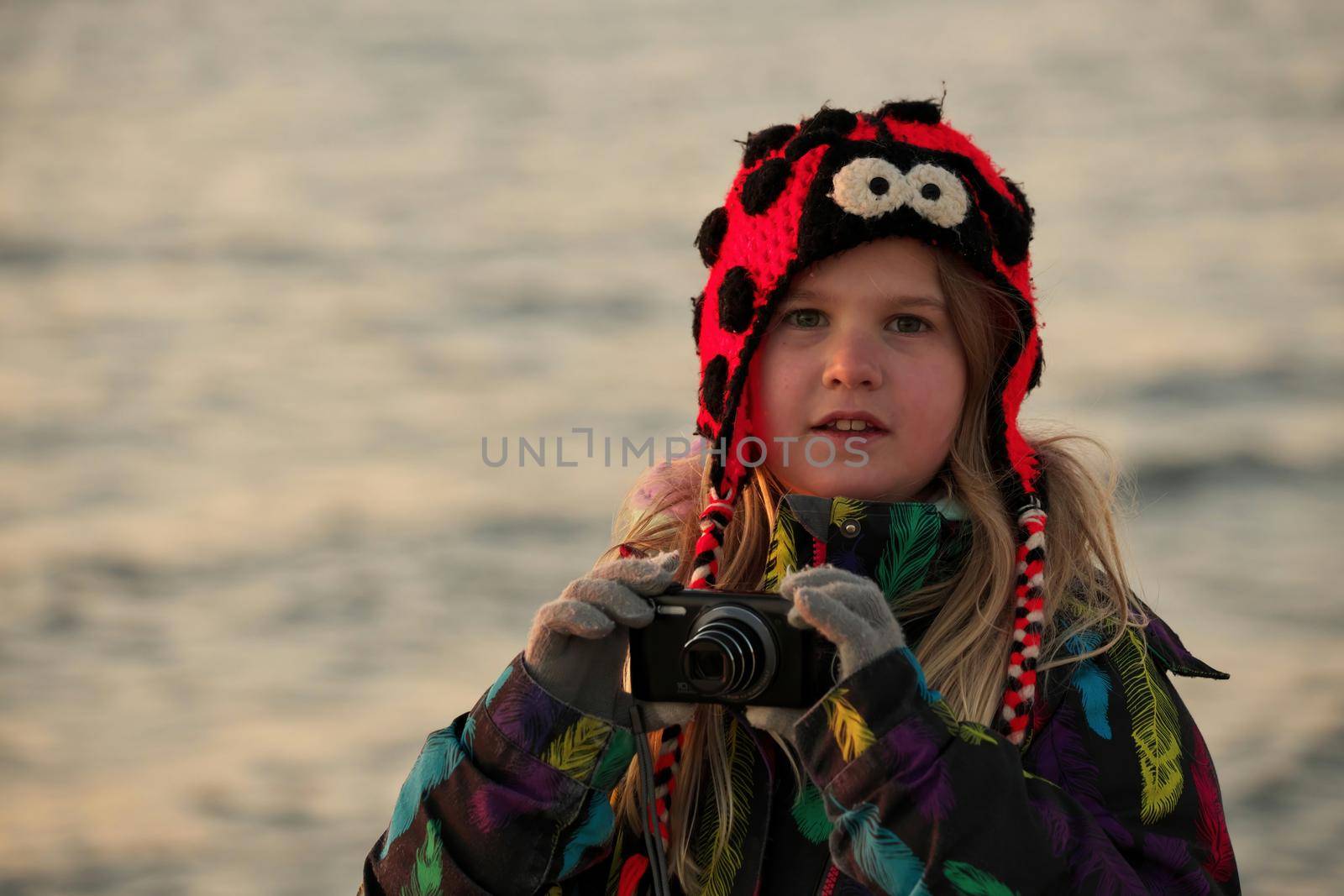 A 9 year old budding photographer practices her camera skills at sunset at the beach. It is cold and she wears a winter jacket and ladybug toque. High quality photo