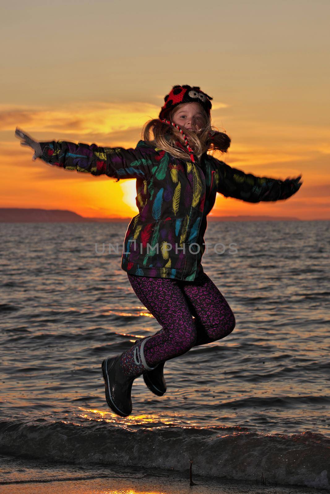 A young girl jumps for joy in front of the setting sun at the beach by markvandam