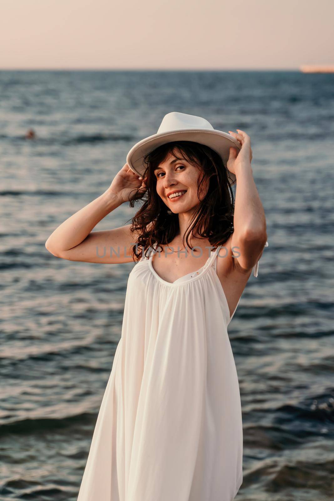 A woman in a white dress and hat is standing on the beach enjoying the sea. Happy summer holidays by Matiunina