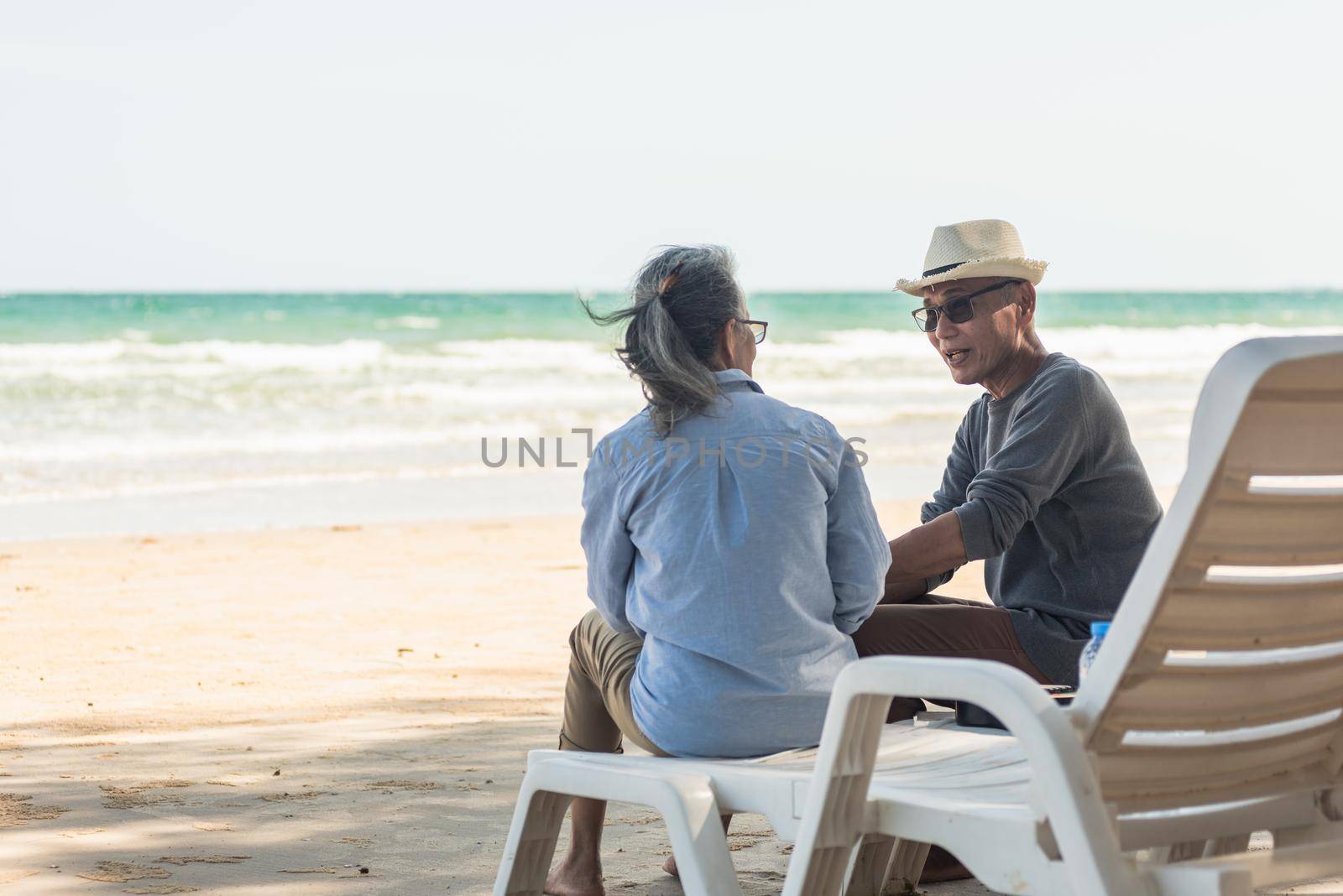 Happy Asian family, senior couple sitting on chairs with backs on beach travel vacation talking together, Romantic elderly enjoy Travel summer vacation, plan life insurance retirement couple concept