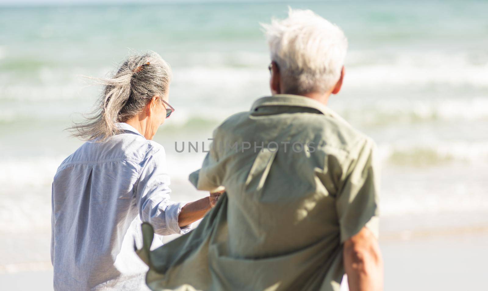 senior man and woman couple holding hands walking to the beach sunny with bright blue sky by Sorapop