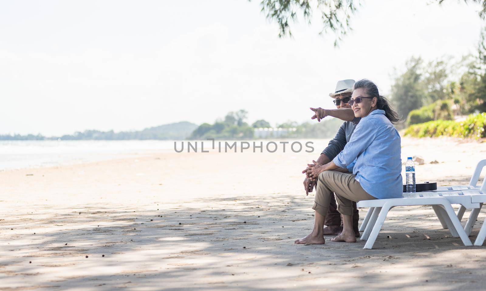 Happy Asian family, senior couple sitting on chairs with backs on beach travel vacation talking together, Romantic elderly enjoy Travel summer vacation, plan life insurance retirement couple concept