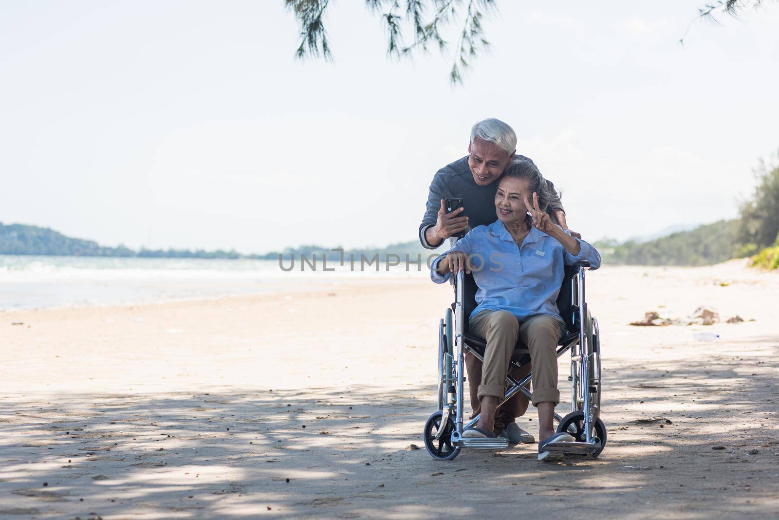 Happy Asian elderly woman sitting in wheelchair and husband is a wheelchair user smartphone taking selfie on the beach, summer vacation, Retirement couple concept