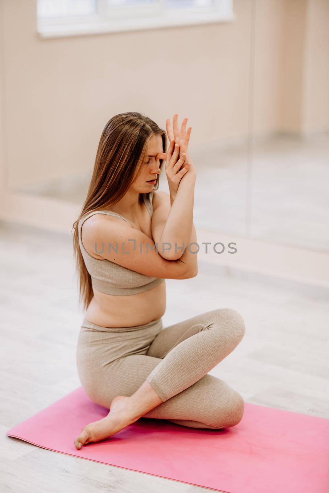Young woman doing yoga in the gym. A girl with long hair and in a beige tracksuit stands in a cow pose on a pink carpet. A woman performs Gomukhasana