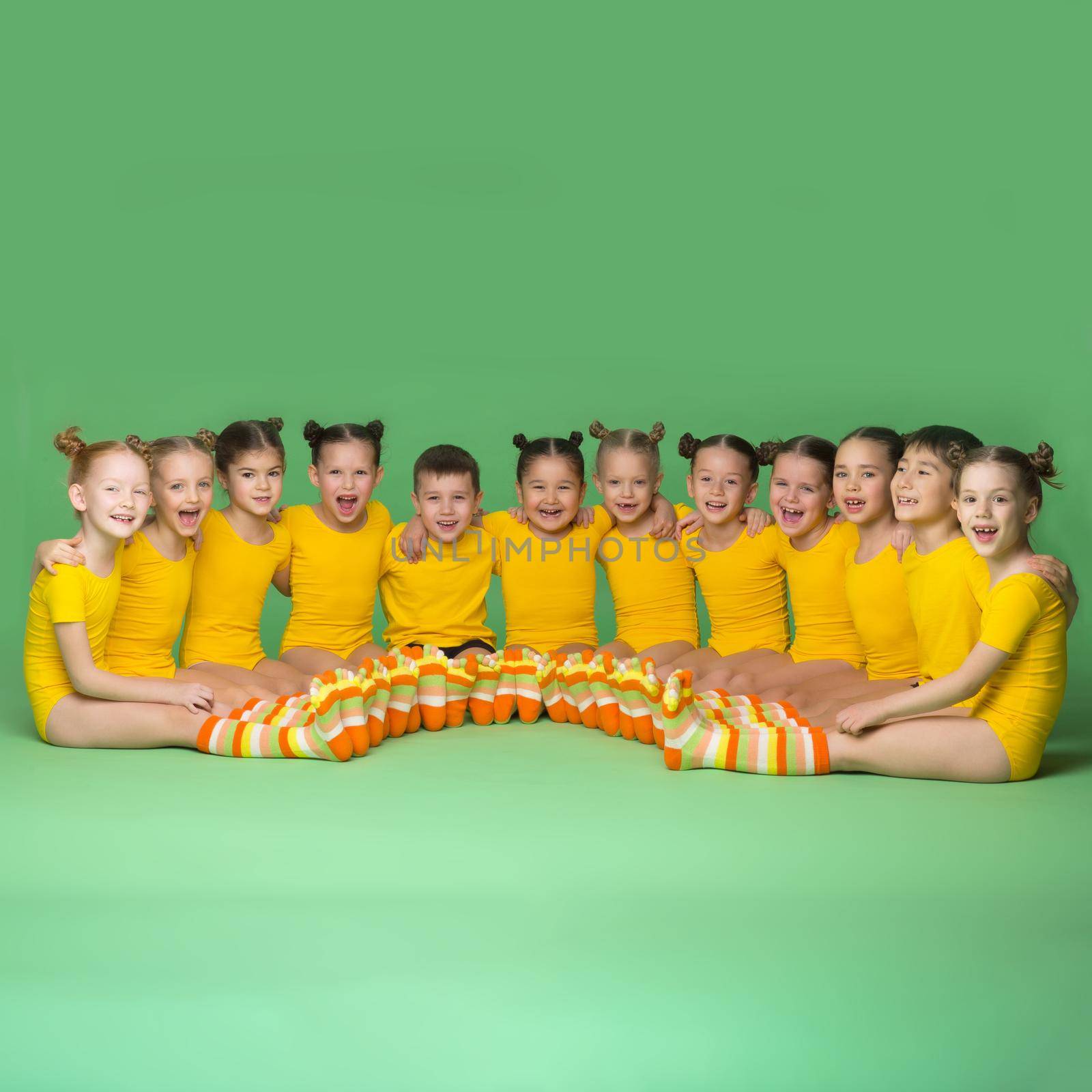 Little children dancers sitting on floor in semicircle. Cute cheerful kids in yellow leotards posing together against green background in studio