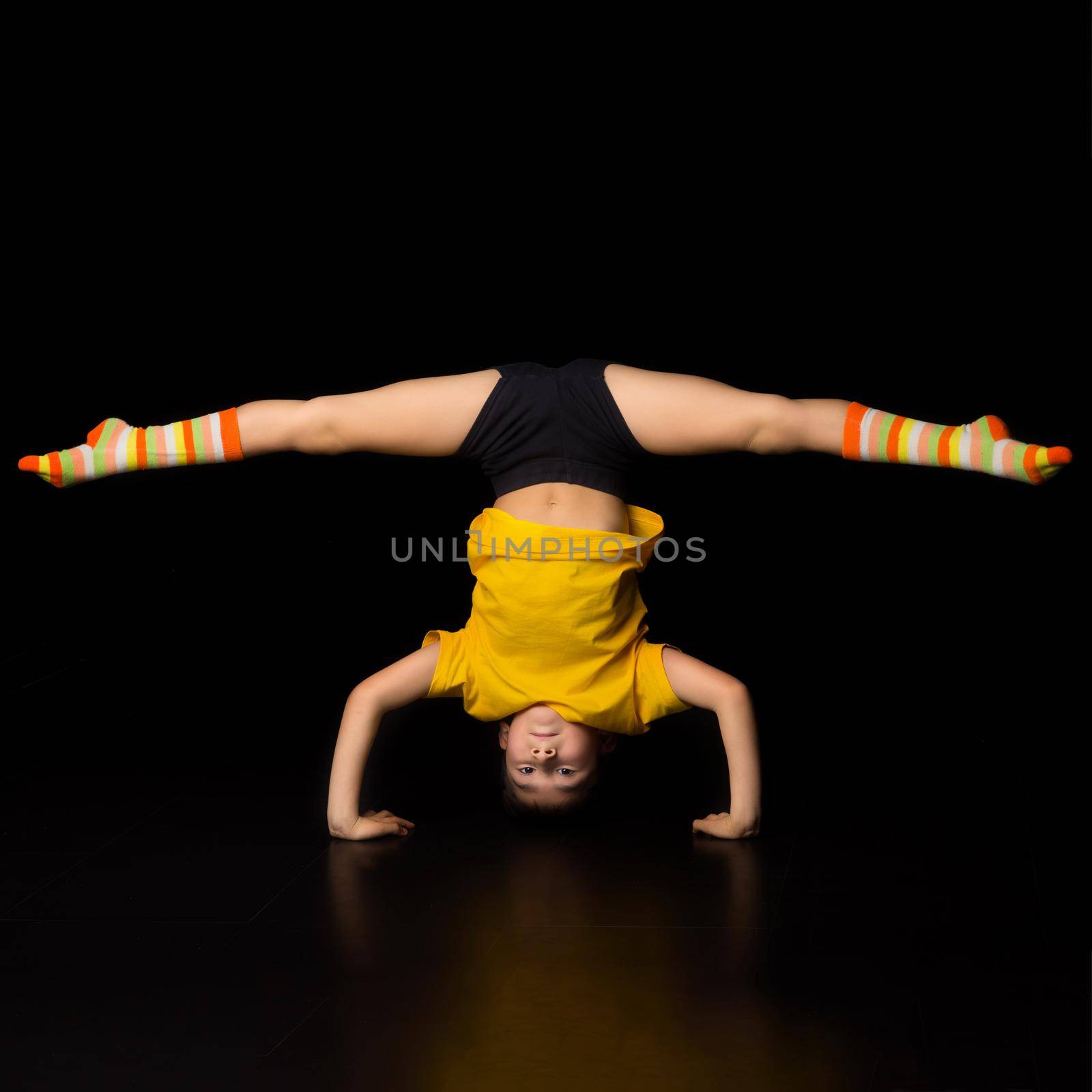 Young boy doing handstand and splits acrobatic element. Boy gymnast or dancer wearing sportswear and striped socks performing gymnastic exercise against isolated black background in studio
