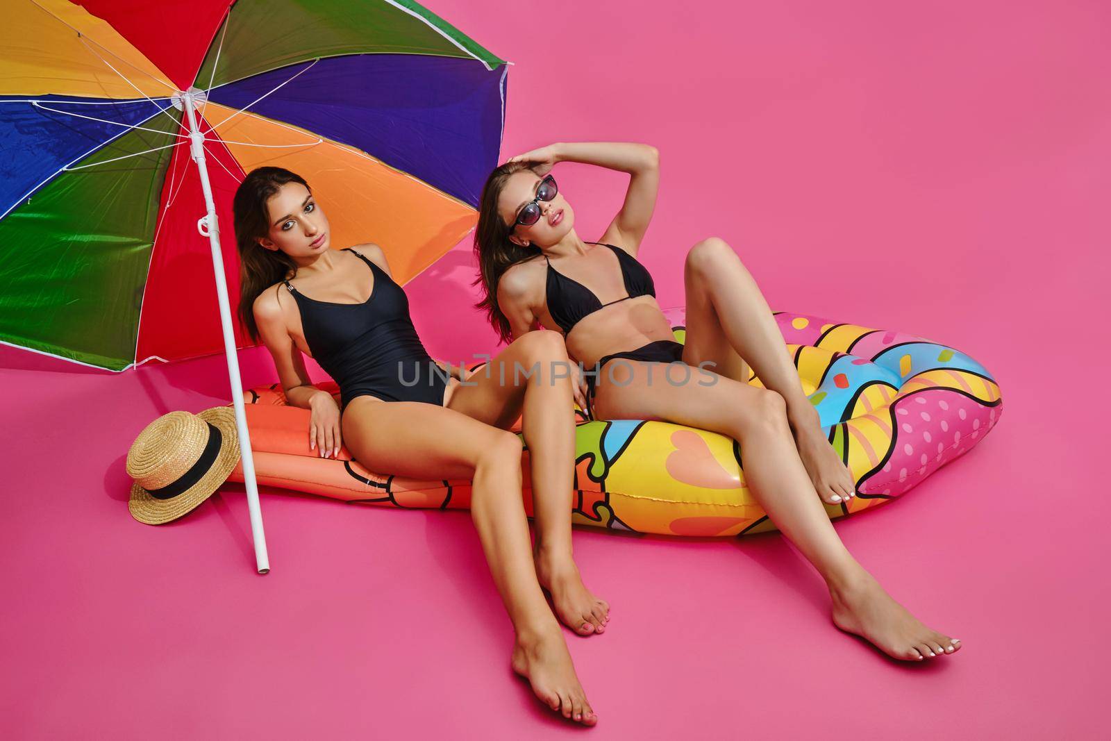 Two attractive carefree girls wearing black bathing suits sitting on colorful air floating mattress under large sun umbrella. Dreams of beach vacation concept. Studio portrait on pink background
