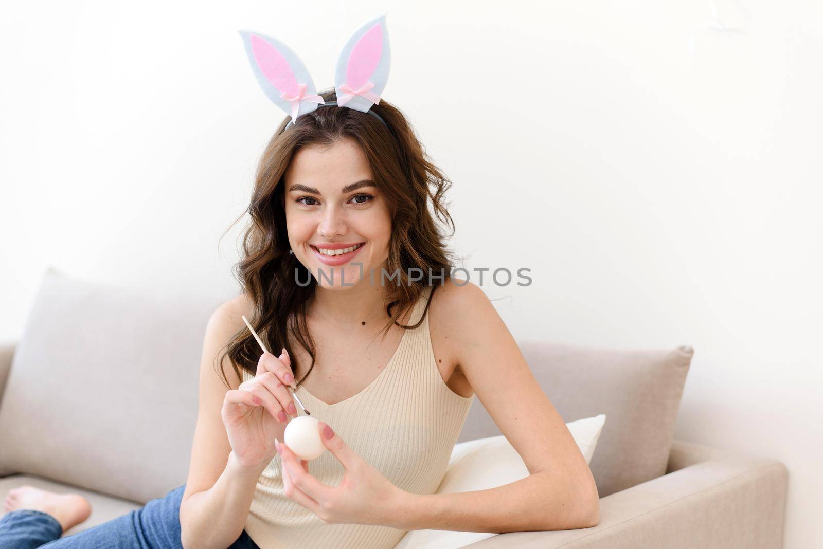 Beautiful young woman in bunny ears paints eggs for easter and looks at the camera