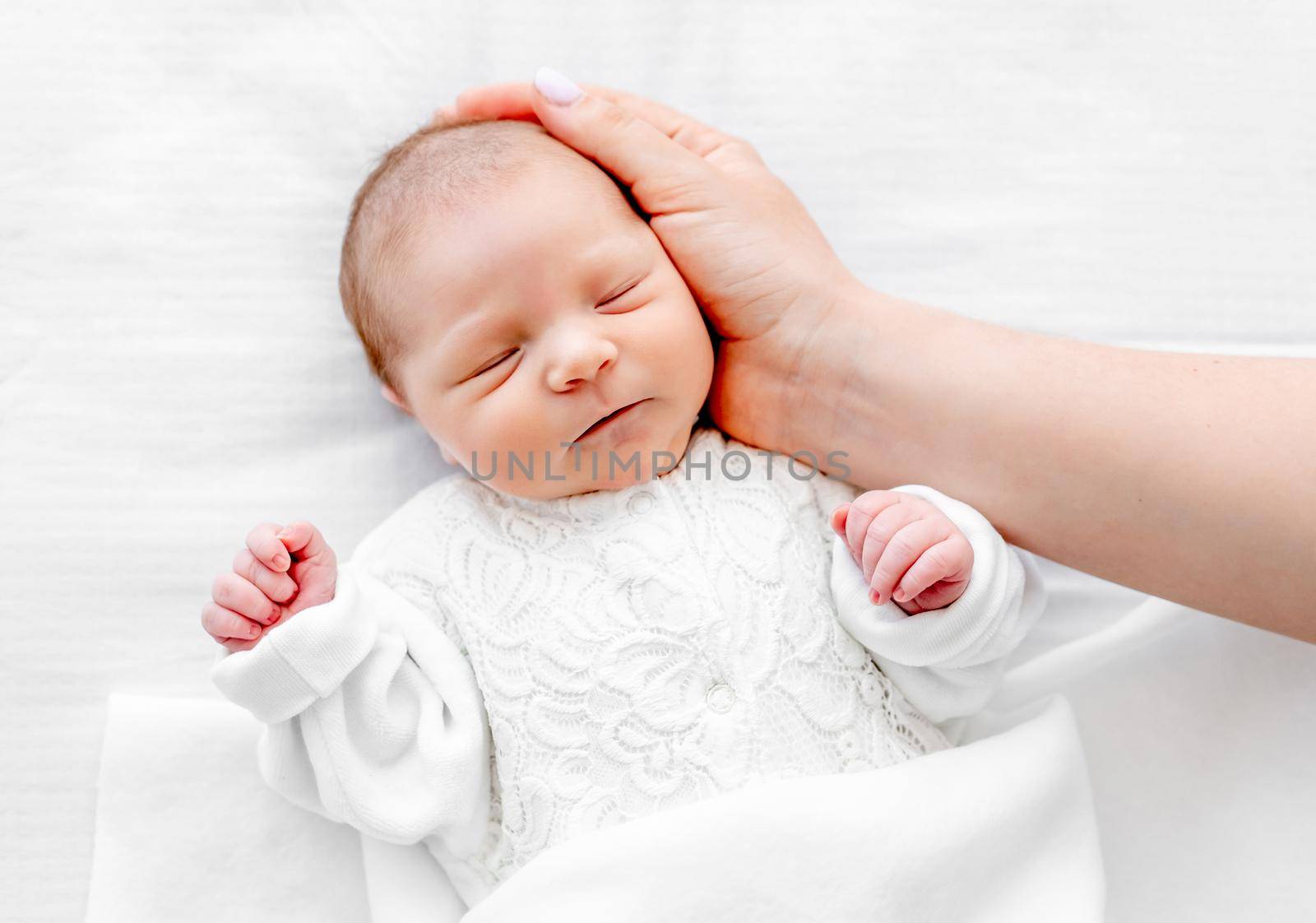 Adorable newborn baby girl wearing white costume lying in the bed, sleeping and her mother hand cares about her. Portrait of cute napping infant child and parent love