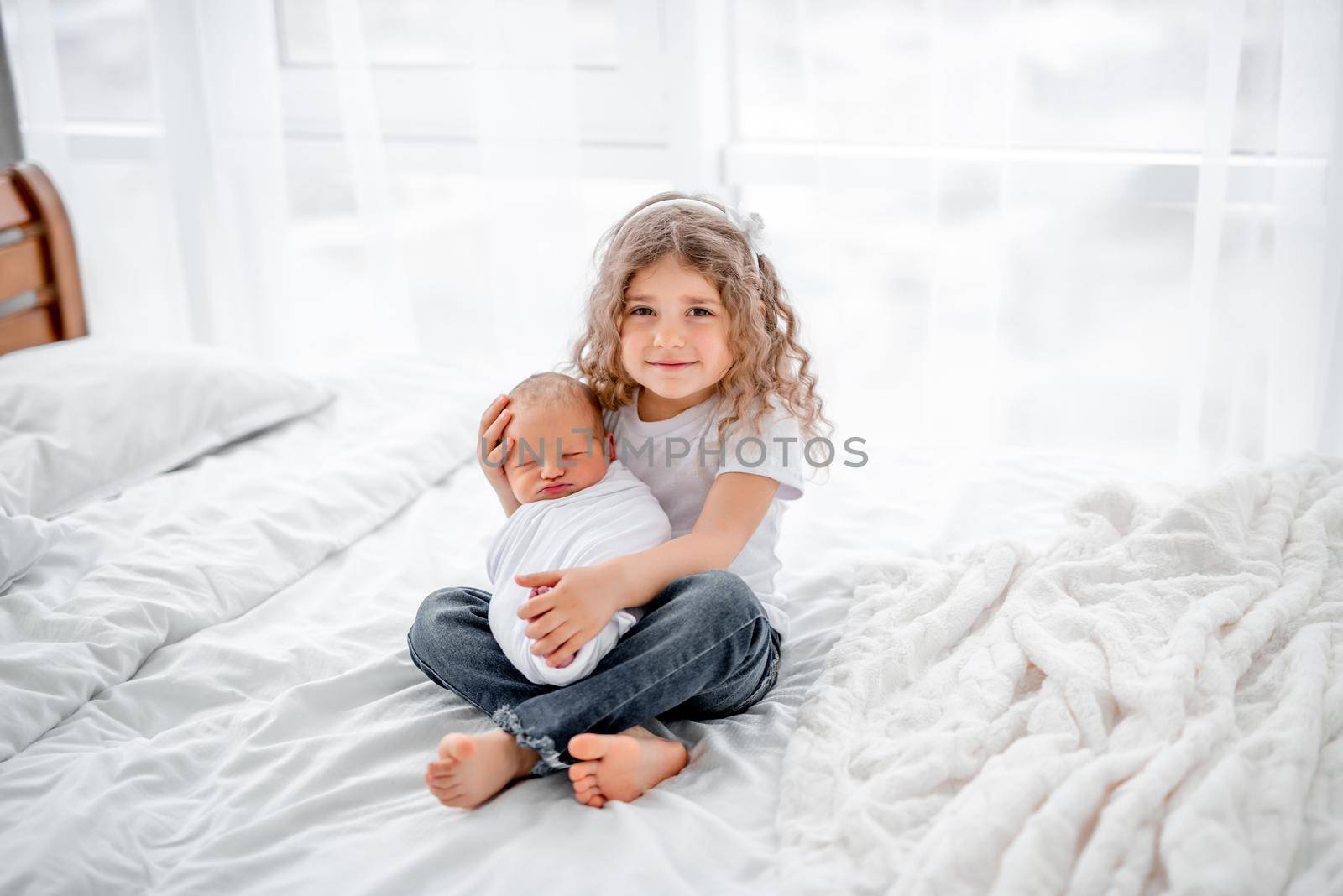 Cute little curly hair girl sitting on the bed and holding adorable newborn baby. Beautiful portrait of sister and her sibling infant child together at home. Happy family moments with children