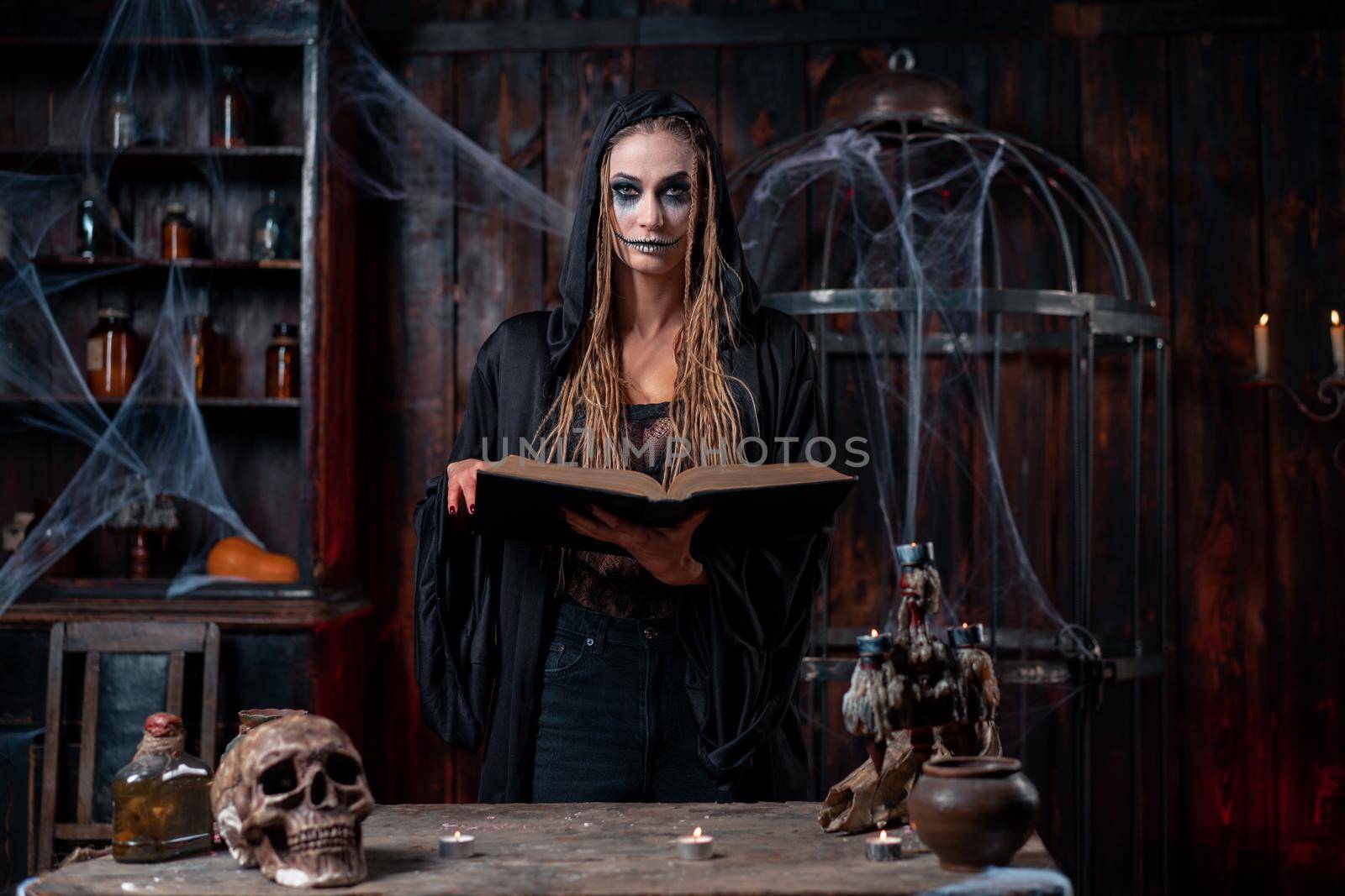 Halloween concept. Witch dressed black hood standing dark spooky room use magic book for conjuring magic spell. Female necromancer wizard gothic interior with skull, cage, spider web