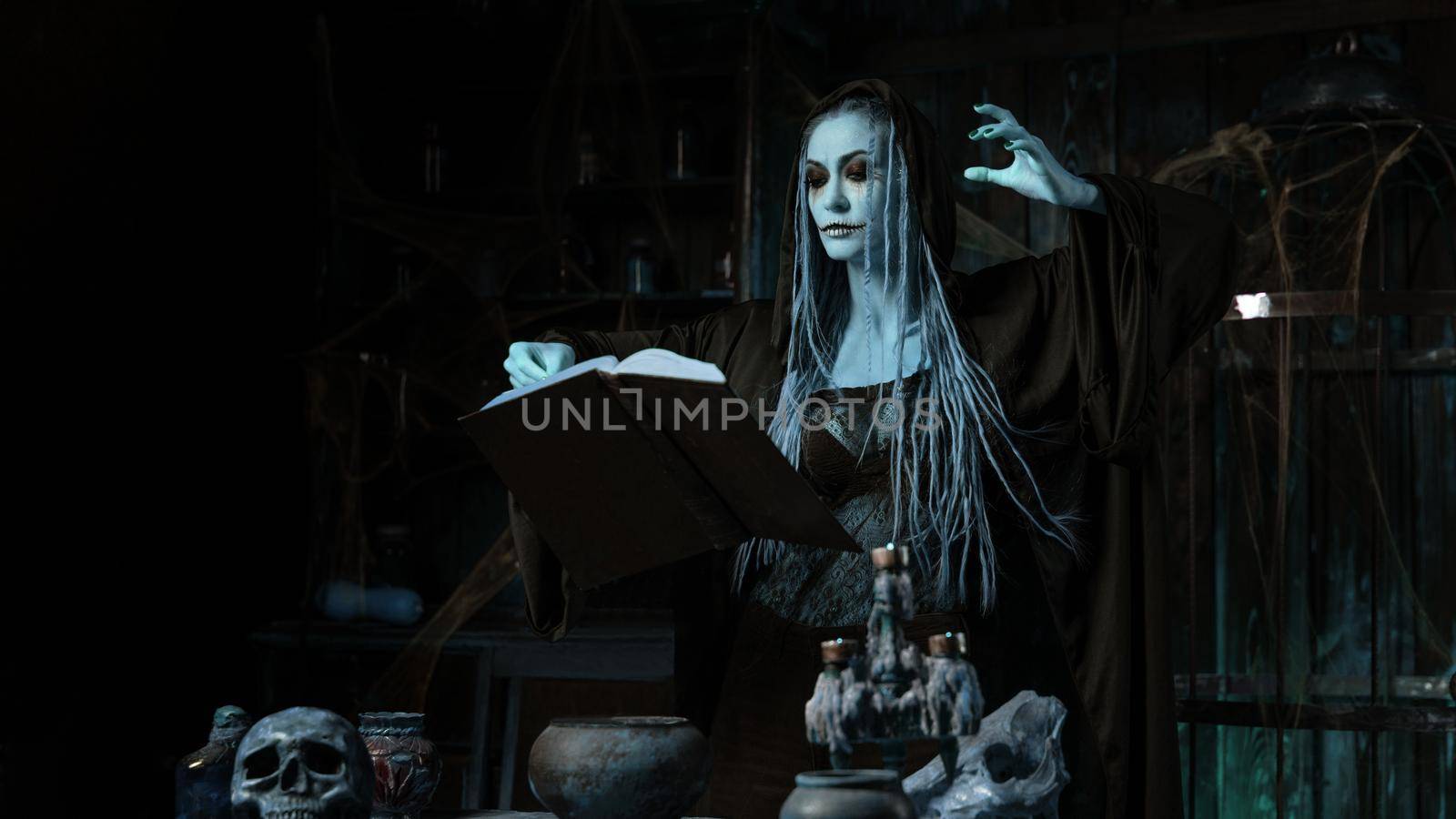 Halloween concept. Dead Cold Blue Witch dressed black hood with dreadlocks standing dark dungeon room use magic book for conjuring magic spell. Female necromancer wizard gothic interior