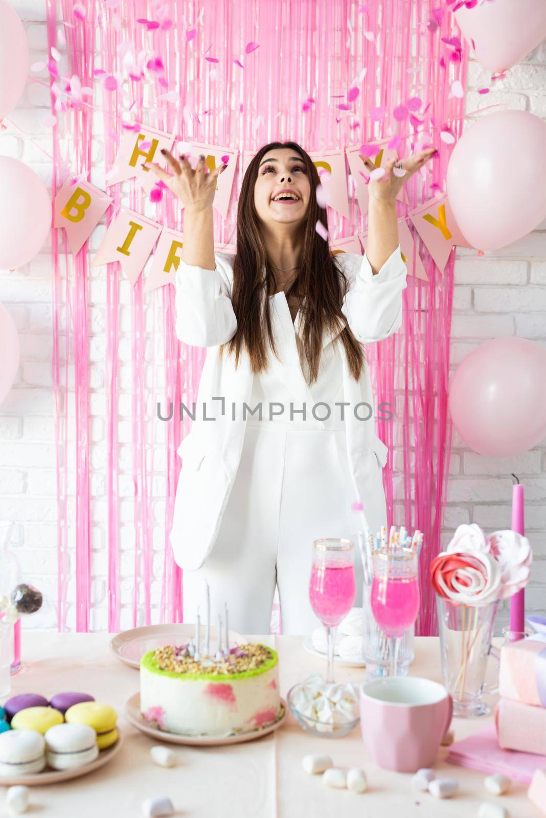 Birthday party. Birthday tables. Attractive brunette woman in white party clothes preparing birthday table with cakes, cakepops, macarons and other sweets, throwing pink confetti