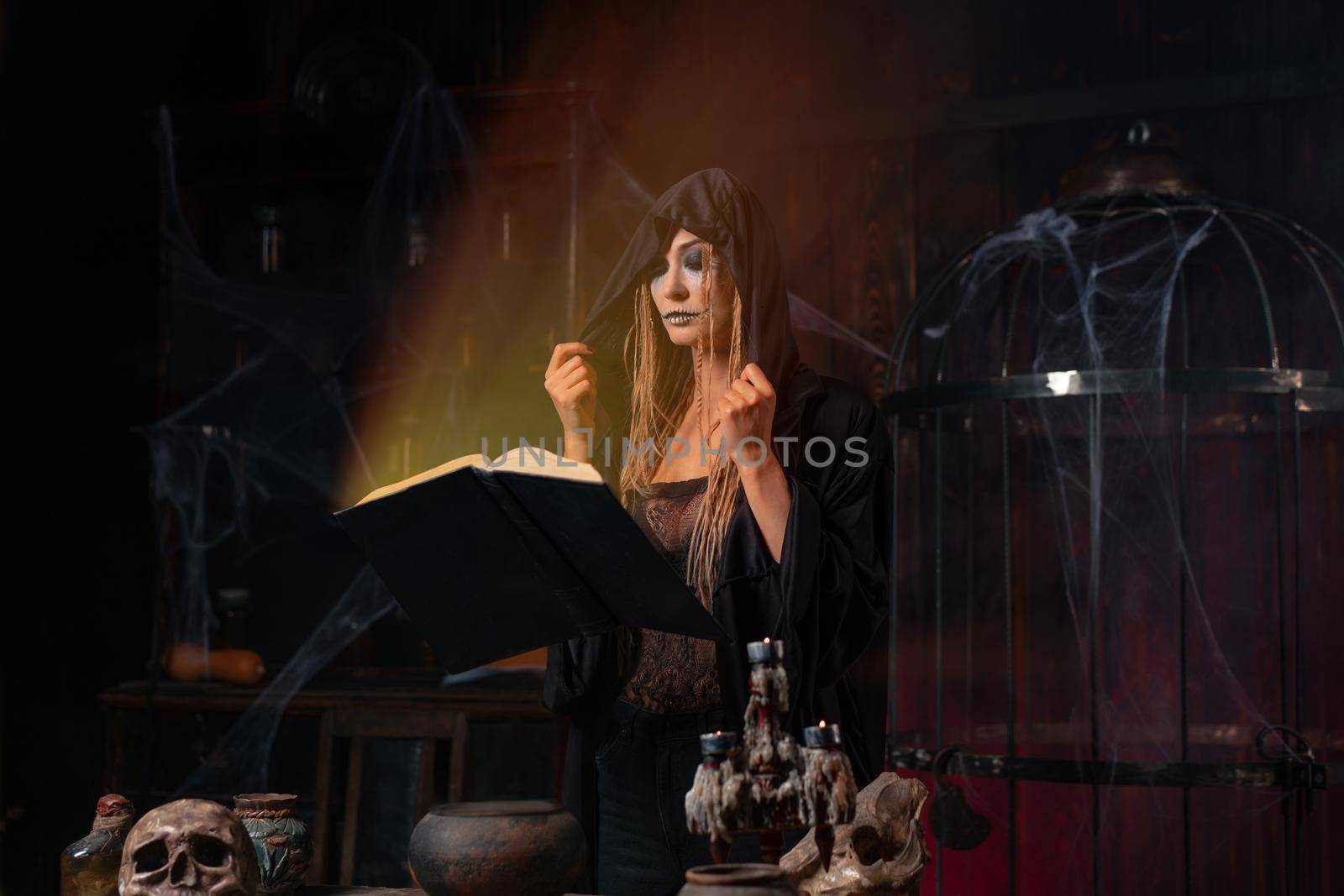 Halloween concept. Witch dressed black hood standing dark dungeon room use magic book see the light from book conjuring magic spell. Female necromancer wizard gothic room with skull, cage, spider web