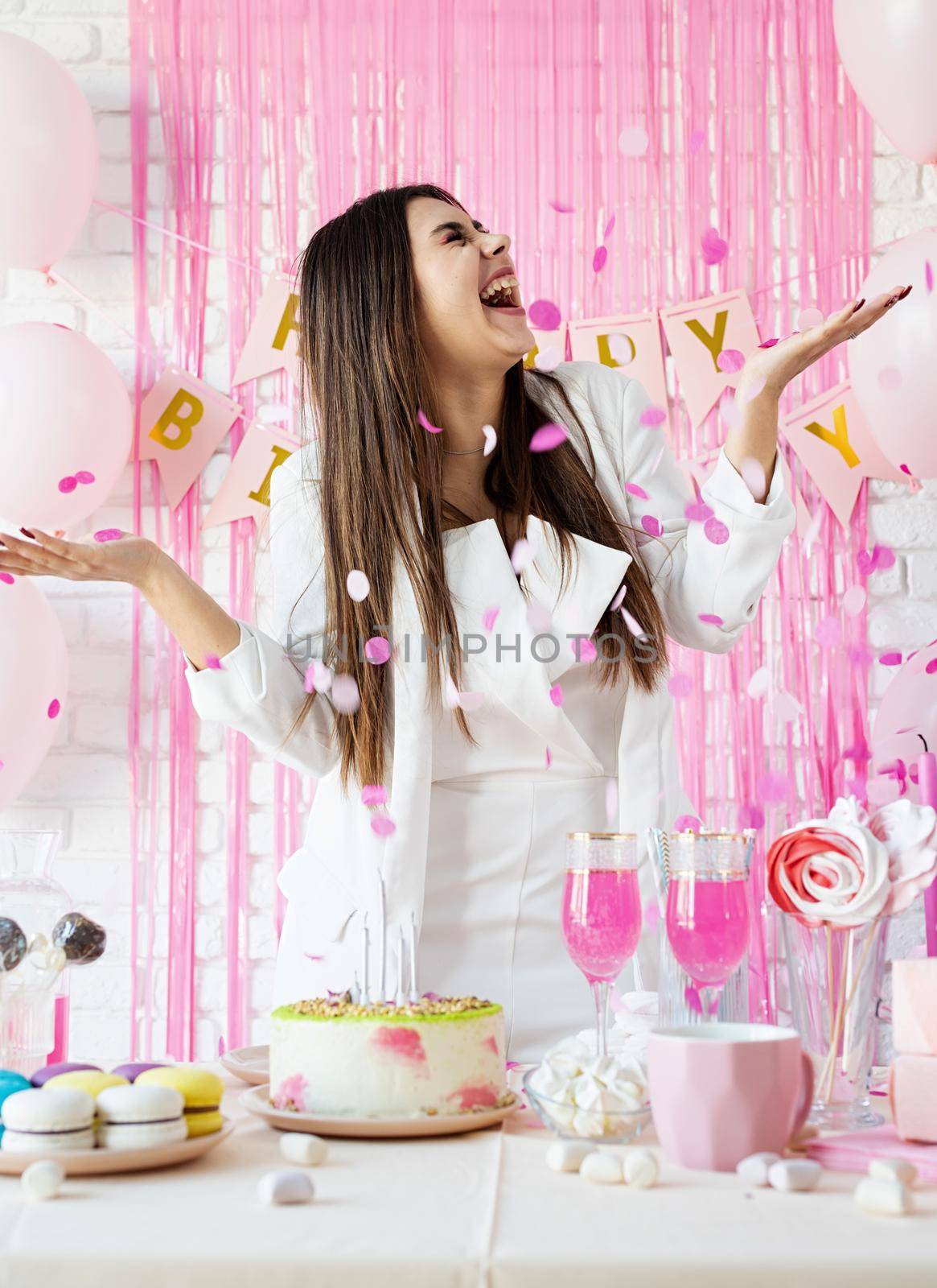 Birthday party. Birthday tables. Attractive brunette woman in white party clothes preparing birthday table with cakes, cakepops, macarons and other sweets, throwing pink confetti