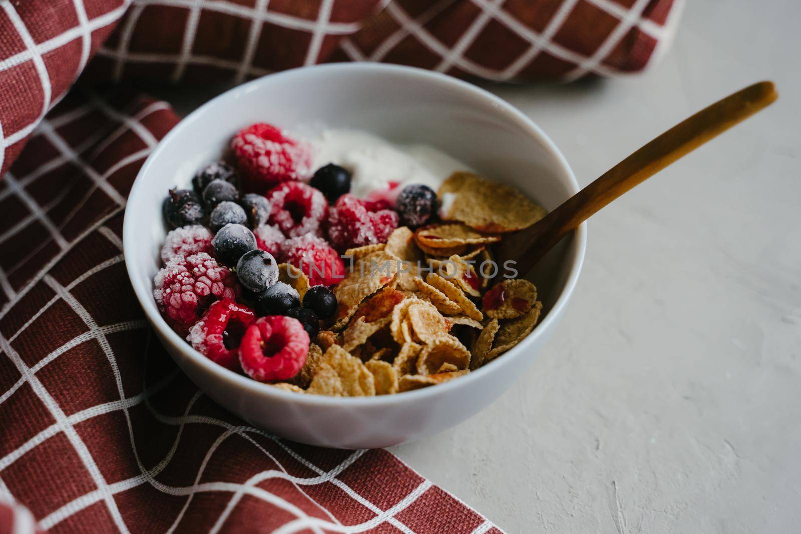 Healthy breakfast with yoghurt, berries and cereals. Wooden spoon. Multi-lacquered cereals for breakfast. Red plaid towel.