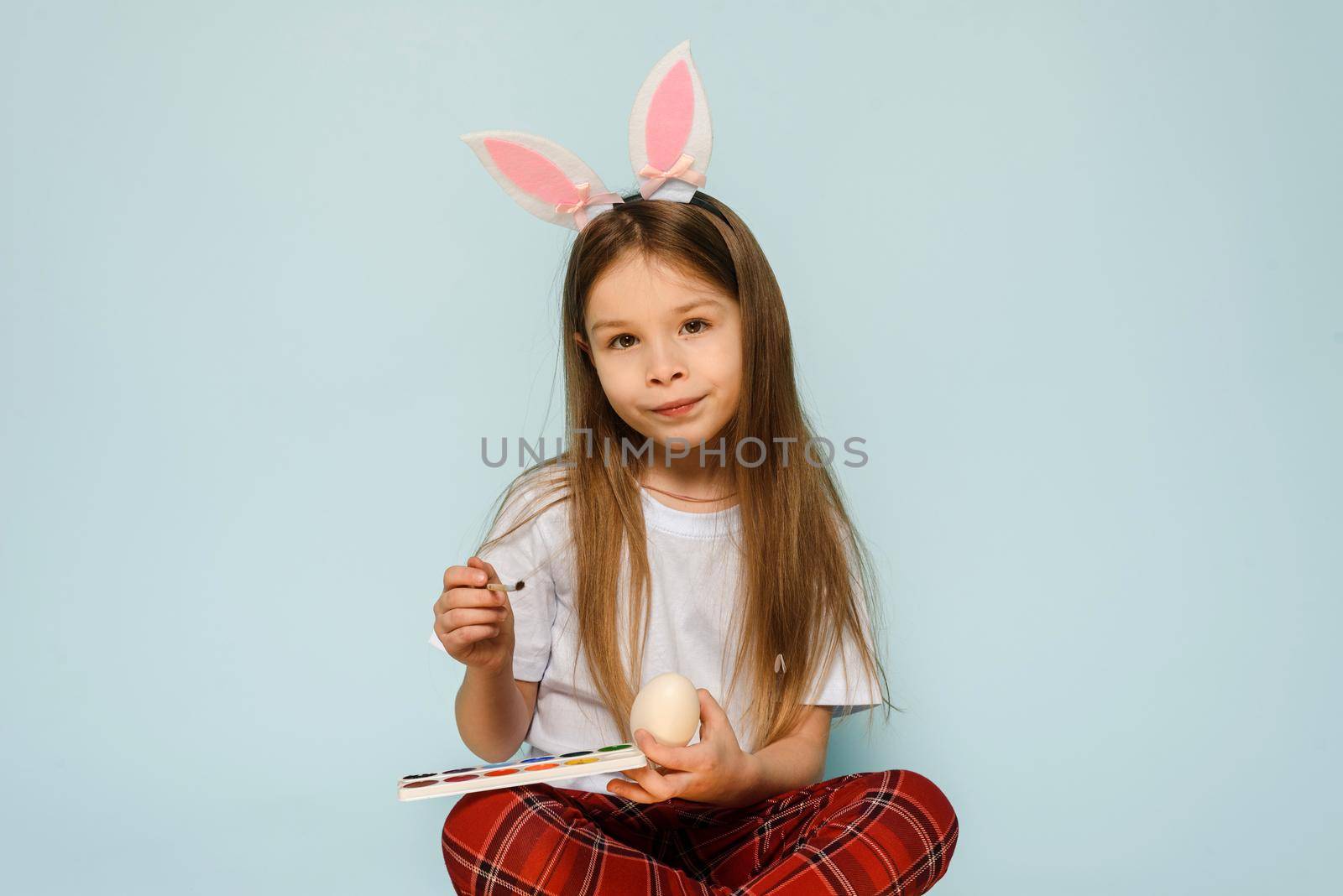 A little girl in rabbit ears looks at the camera and paints eggs for Easter while sitting in the studio. The concept of traditions and preparation for the holiday