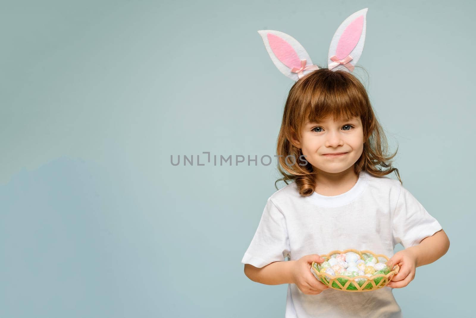 A cheerful girl with rabbit ears on her head with a basket of colored eggs in her hands on a blue background. Easter concept
