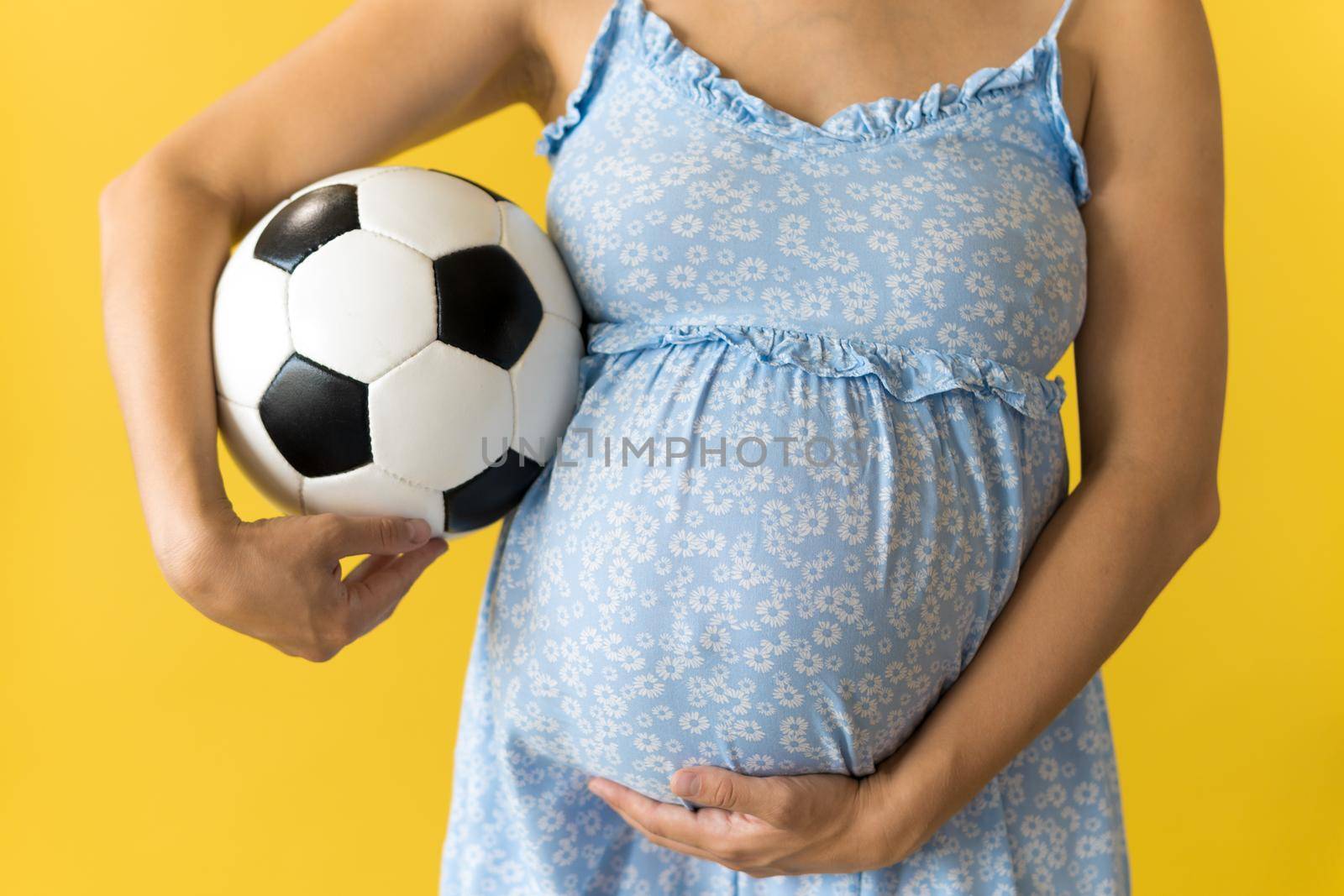 Motherhood, femininity, football, sport, dairy, hot summer. croped unrecognizable pregnant young pretty woman in floral blue dress holds soccer white and black ball rubs tummy on yellow background.