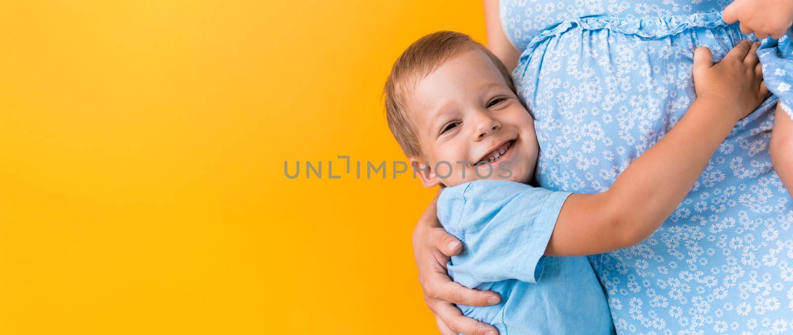 Motherhood, femininity, love, Childhood, hot summer - croped banner pregnant unrecognizable woman blue dress little preschool boy son sibling hug mum big belly brother on yellow background copy space by mytrykau