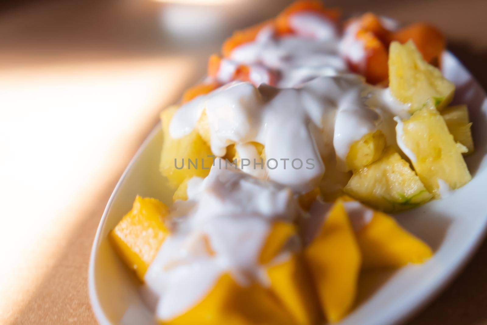 Close-up of plate of sliced mango, pineapple, and papaya, with cream on top. Tasty creamy dessert with fresh fruit slices above brown table. Healthy food and breakfast