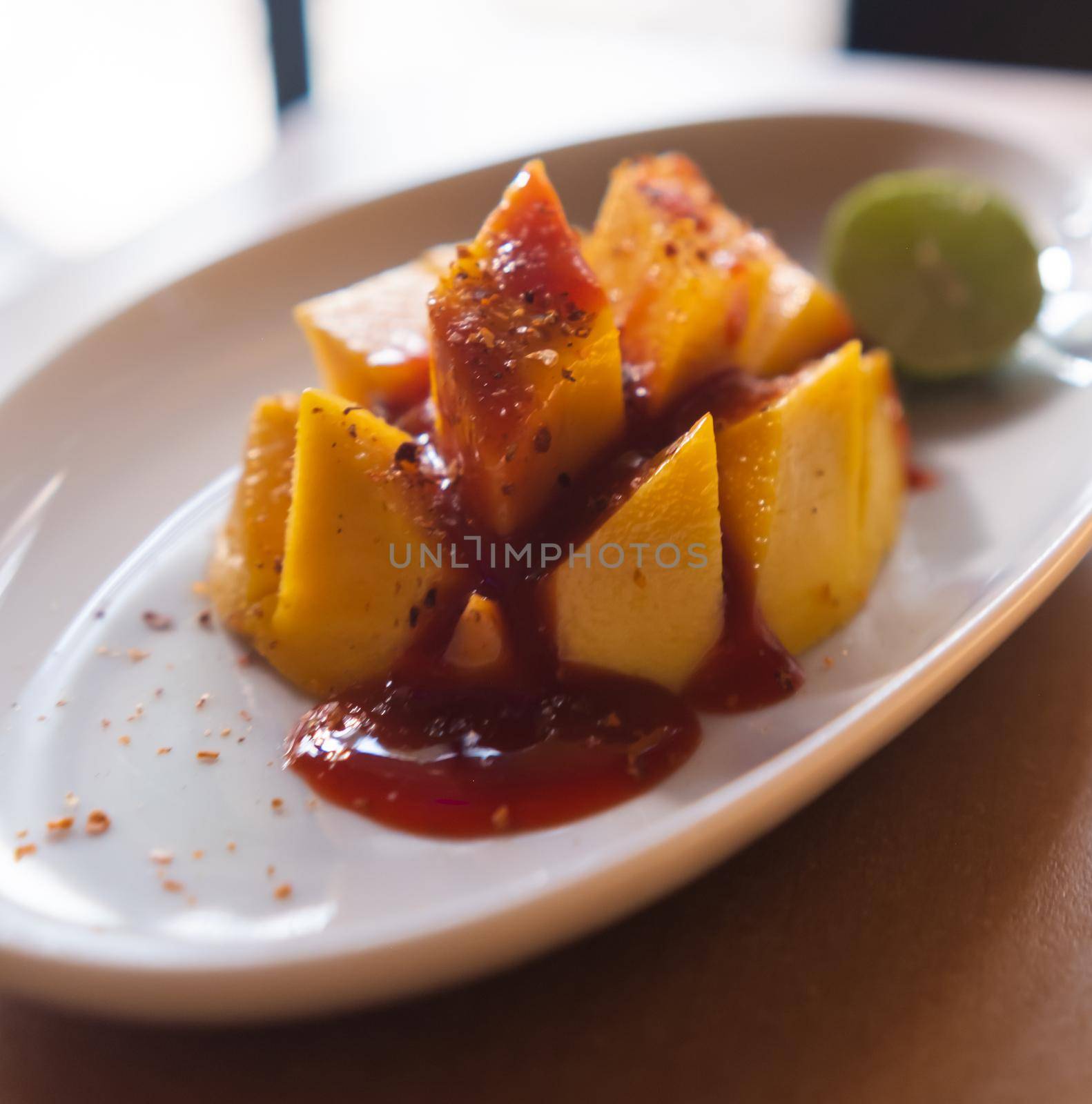 Close-up of white plate of chopped mango with chili powder and Mexican chamoy sauce. Fresh tropical fruit slices with spicy and sour condiment on wooden table. Healthy food and snacks