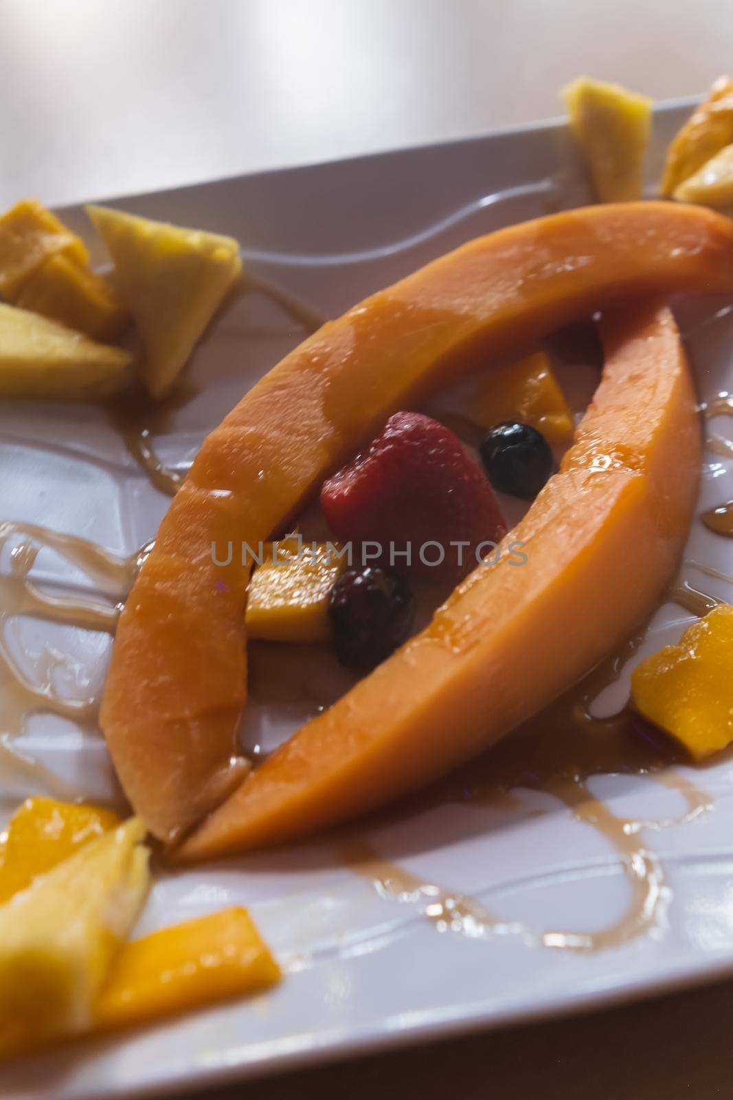 Close-up of papaya and pineapple slices, blueberries, and strawberries on white square plate. Fresh honey and delicious sliced fruit arranged on porcelain plate. Healthy food and snacks