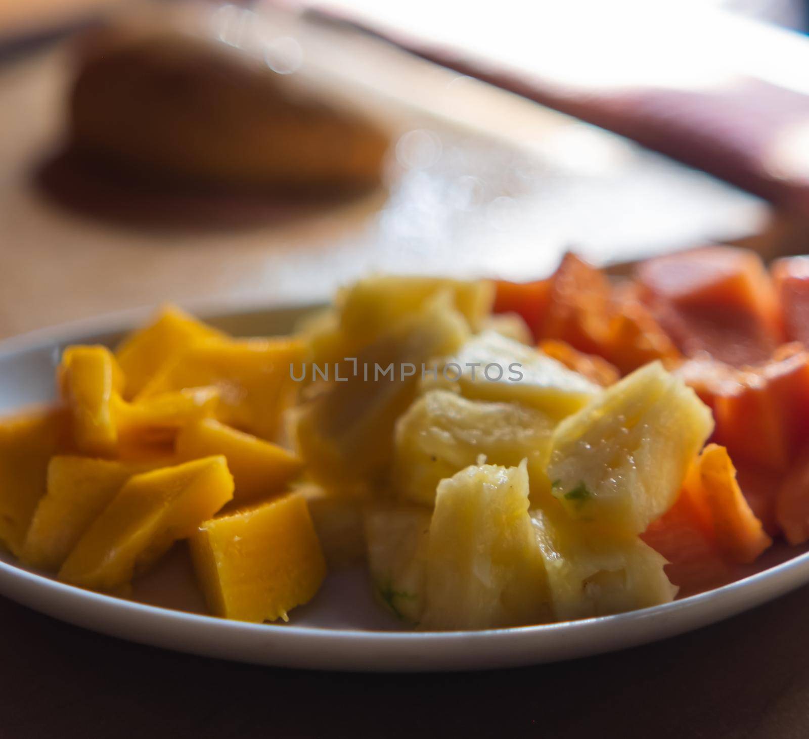 Close-up of white plate of mango, pineapple, and papaya slices on table with blurry and bright background. Tasty and fresh sliced fruit on an oval porcelain plate. Healthy food and breakfast