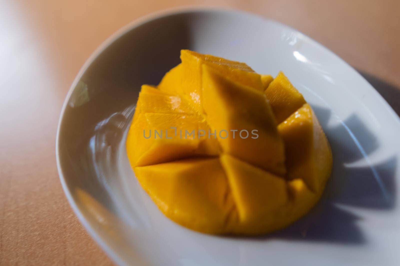 Close-up of diced mango on oval white plate above brown table. Fresh yellow tropical fruit above shiny wooden surface. Healthy snack preparation