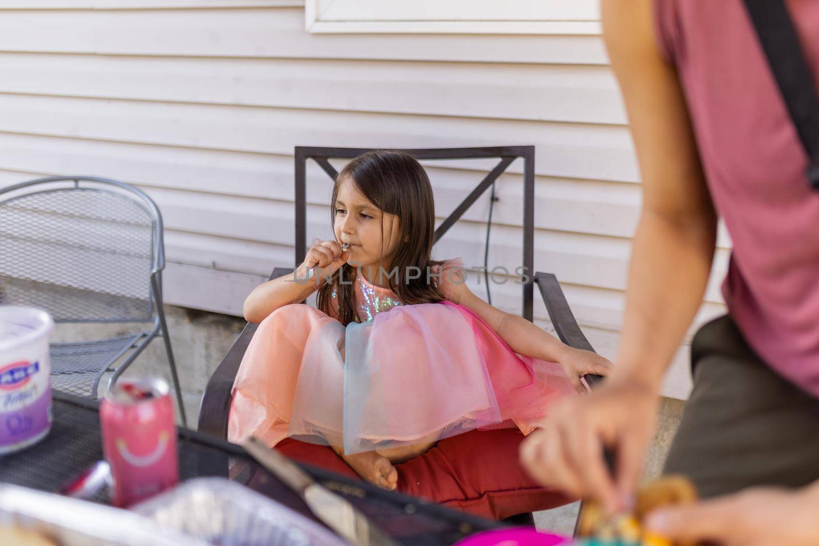 Cute little barefoot girl in pink princess dress sitting at table outside. Adorable young girl on comfortable chair with white wall as background. Kids having fun outdoors