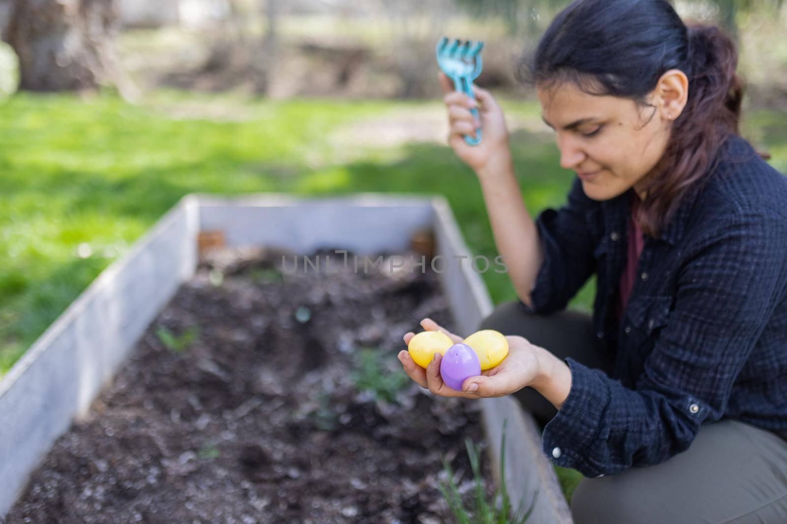 Beautiful brunette woman hiding colorful eggs in empty planter box in yard. Happy young woman holding little plastic rake and plastic eggs above dirt with blurry background. Easter and outdoors