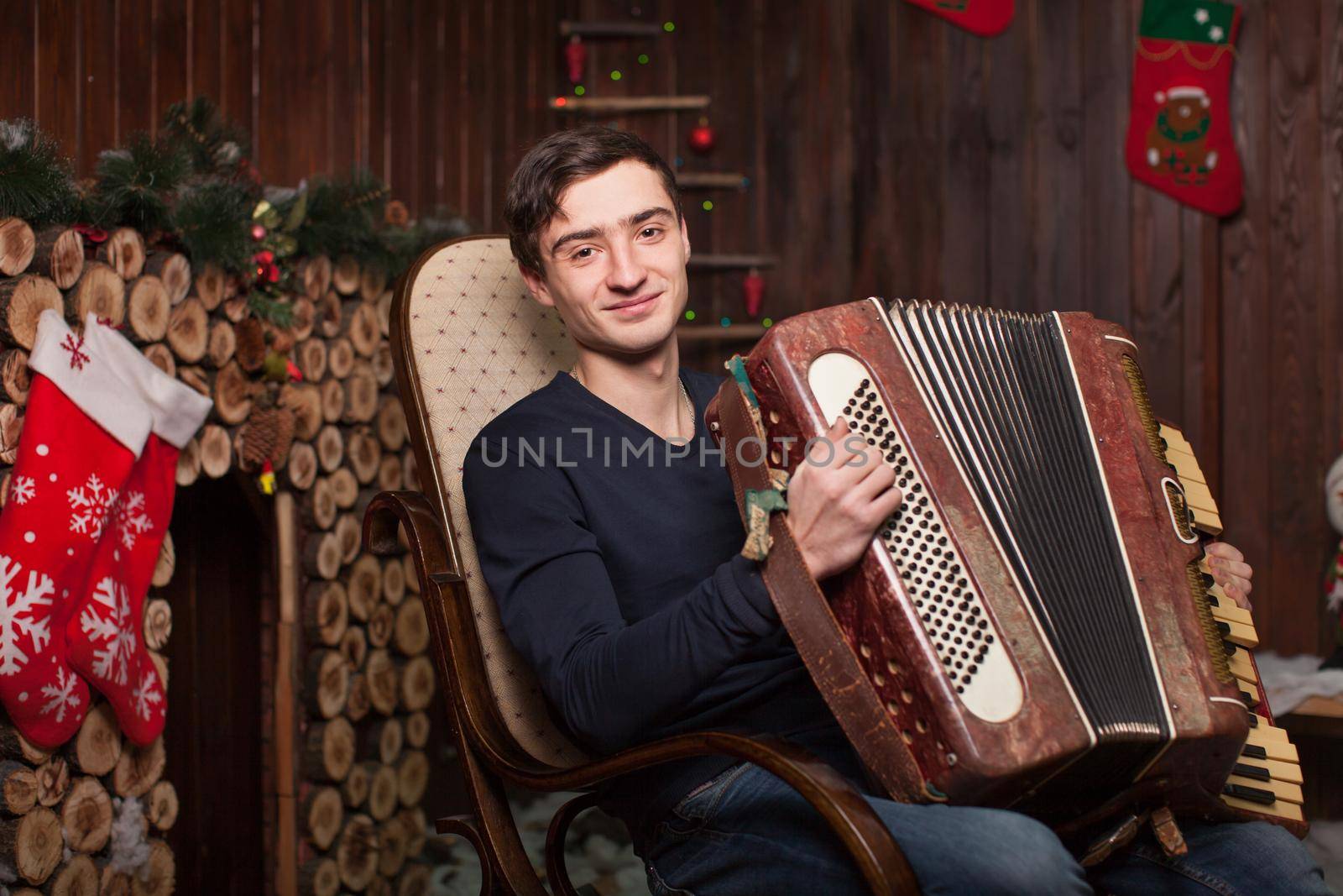A young guy sits on an armchair by the fireplace and plays an accordion