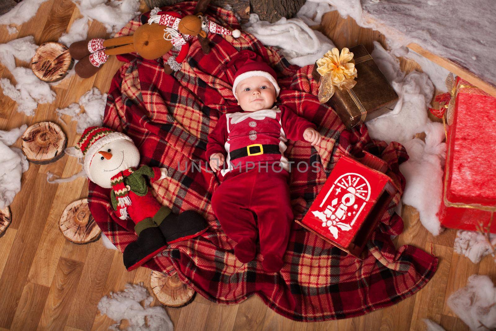 A child in Santa Claus costume rests on a red plaid surrounded by toys