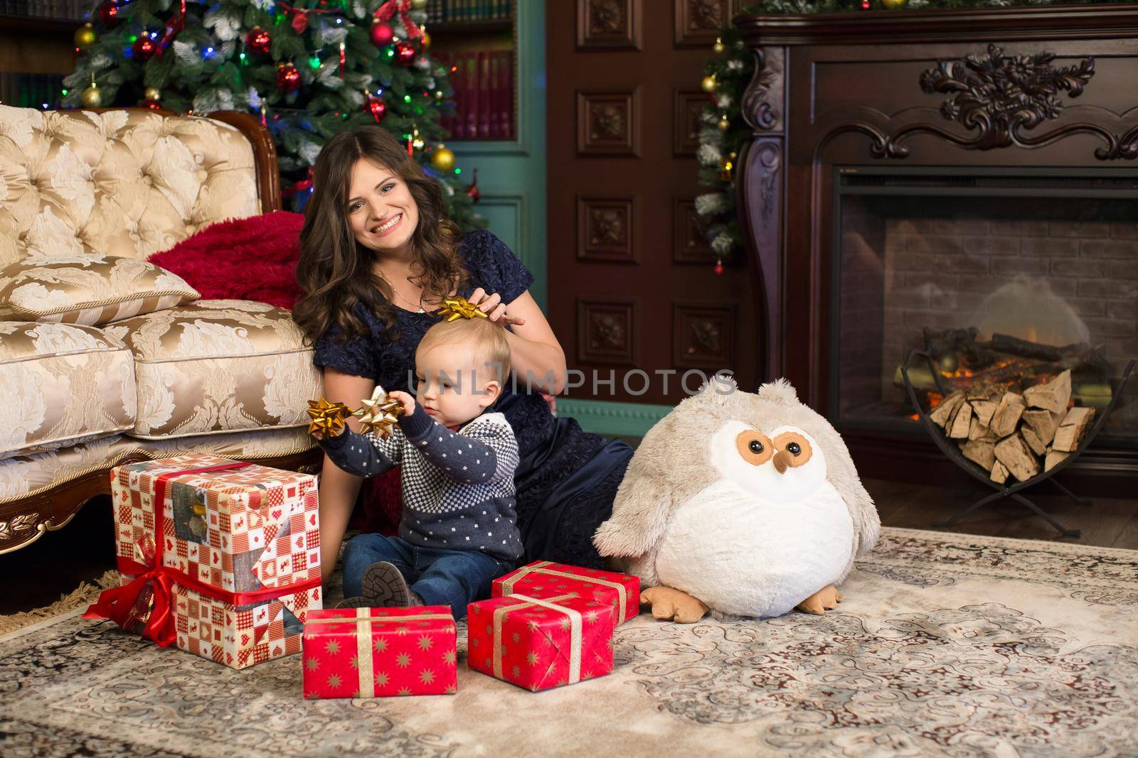 Mother and son open Christmas gifts on the carpet near the Christmas tree and the fireplace.