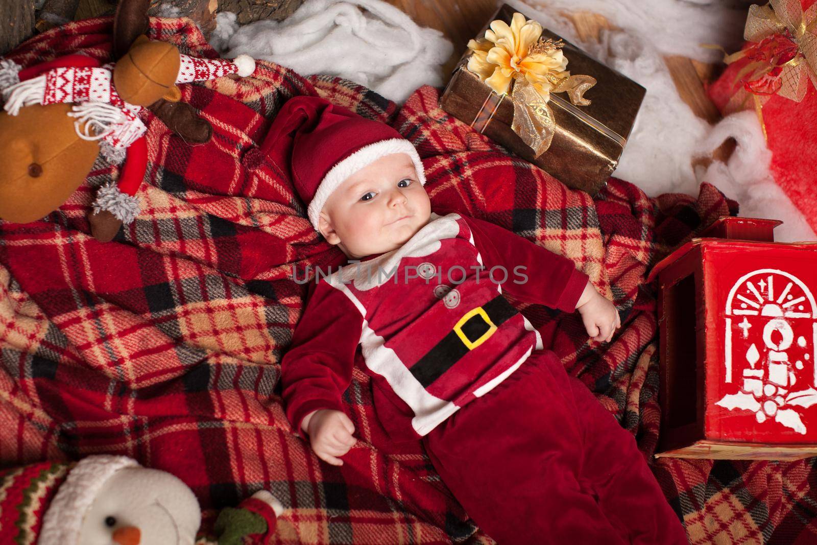 A child in Santa Claus costume rests on a red plaid surrounded by toys
