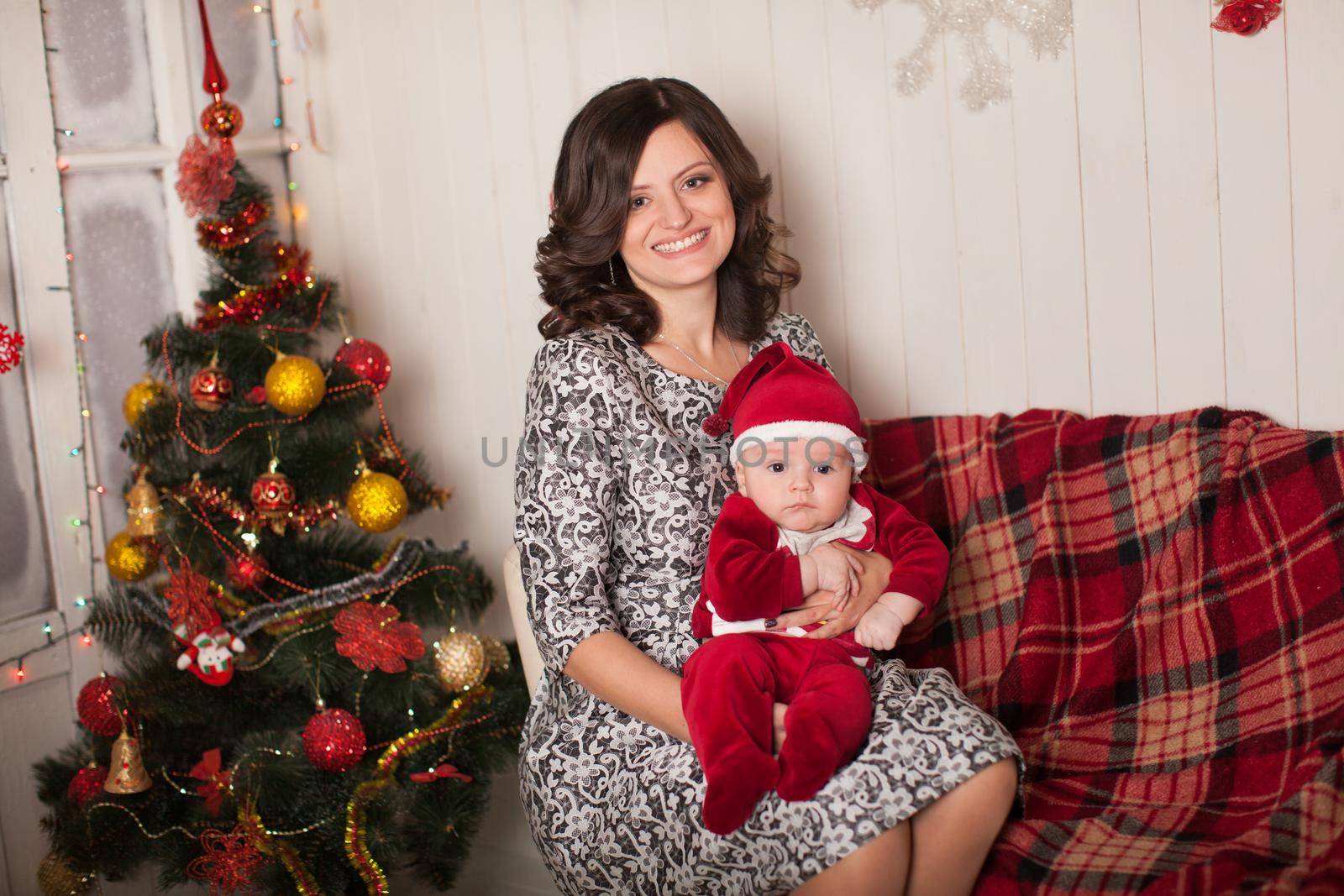 Mom and son in Santa Claus costume in a home setting near a Christmas tree against a white wooden wall