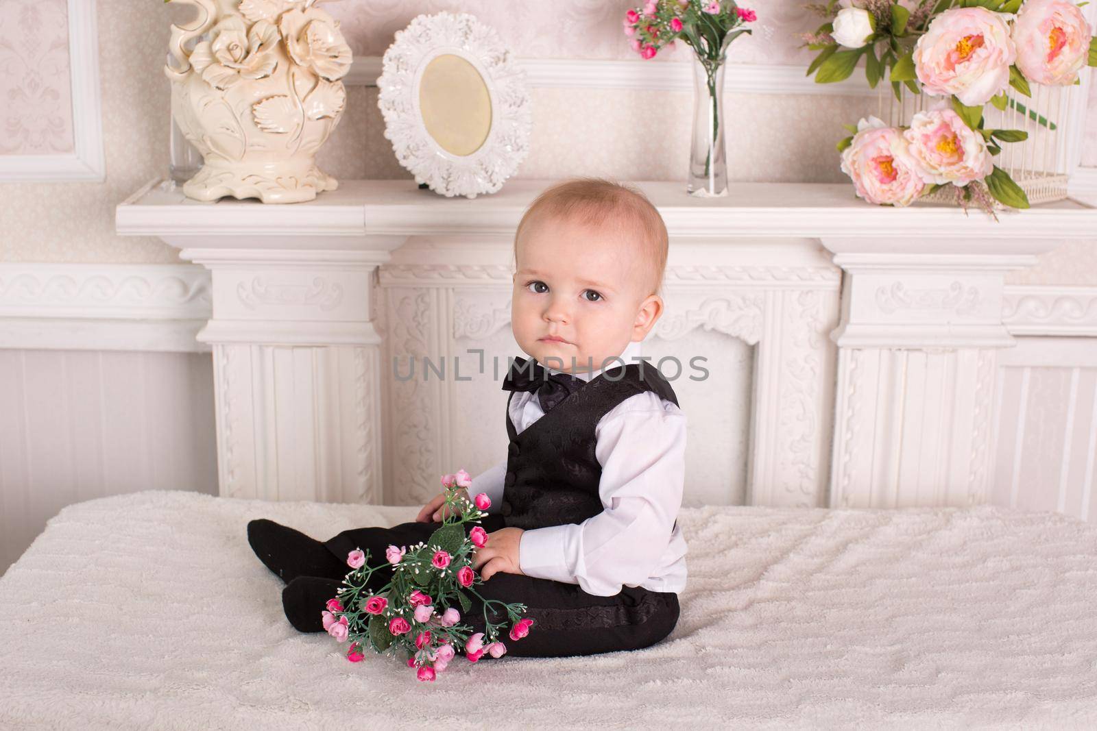 Child in a tuxedo sitting on the bed next to the fireplace with flowers in their hands.